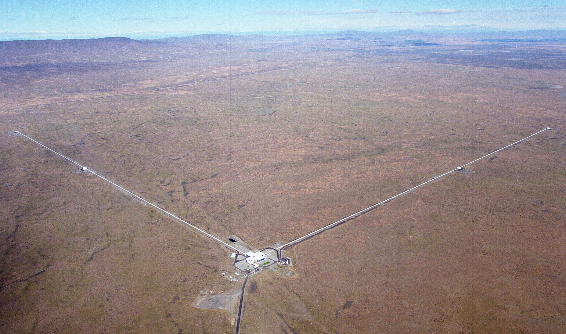 The L-shaped tube of the LIGO detector splayed across a brown landscape with low mountains in the background.