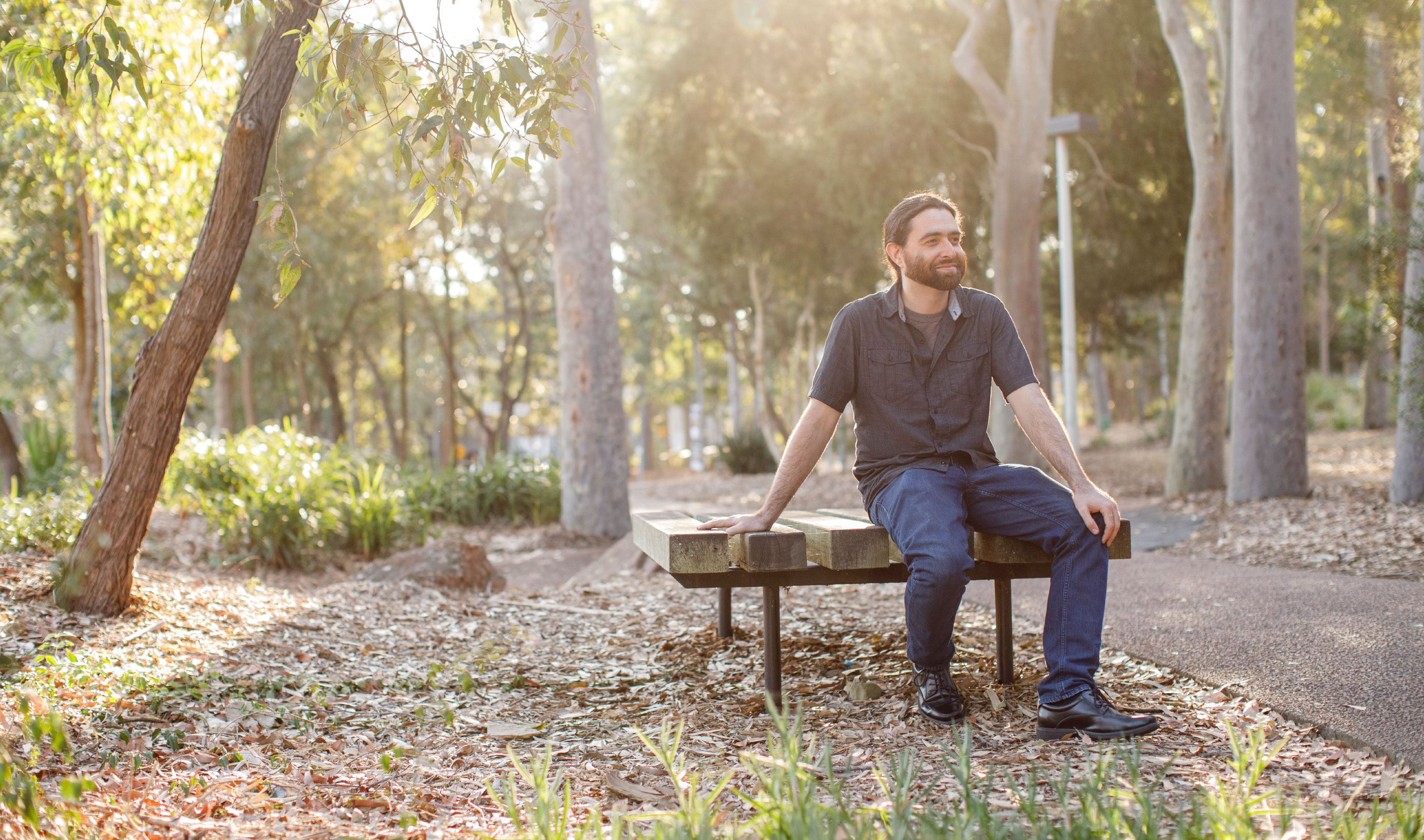 Micheal Assis in jeans and a blue shirt sitting outside on a bench