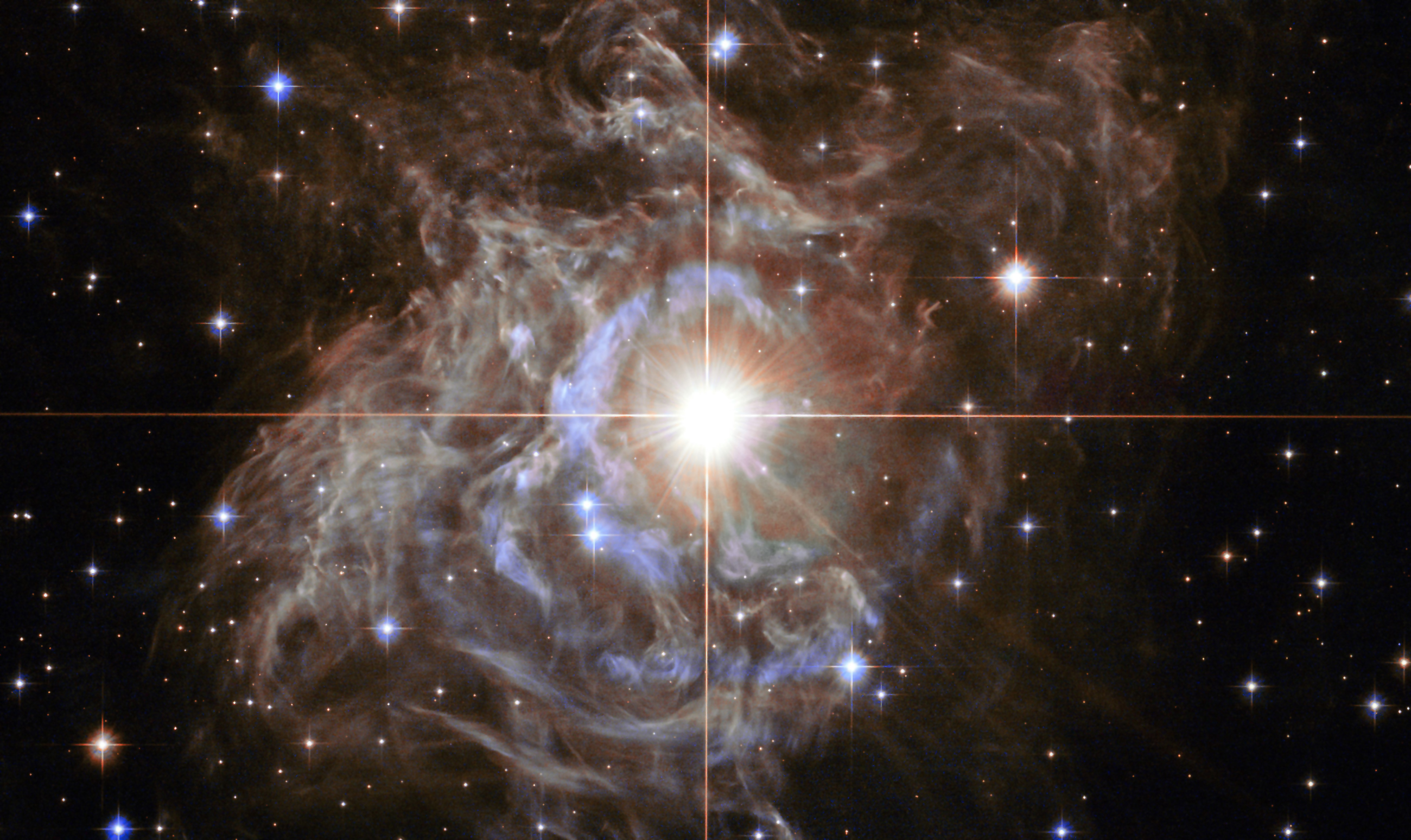 Lede art for "A Radically Conservative Solution for Cosmology’s Biggest Mystery": This Hubble image shows RS Puppis, a type of variable star known as a Cepheid variable. As variable stars go, Cepheids have comparatively long periods— RS Puppis, for example, varies in brightness by almost a factor of five every 40 or so days. RS Puppis is unusual; this variable star is shrouded by thick, dark clouds of dust enabling a phenomenon known as a light echo to be shown with stunning clarity. These Hubble observations show the ethereal object embedded in its dusty environment, set against a dark sky filled with background galaxies.