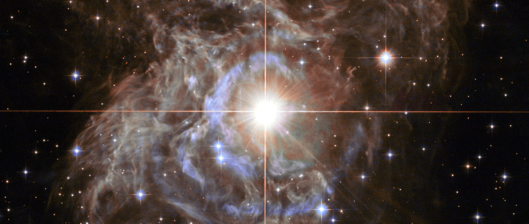 Lede art for "A Radically Conservative Solution for Cosmology’s Biggest Mystery": This Hubble image shows RS Puppis, a type of variable star known as a Cepheid variable. As variable stars go, Cepheids have comparatively long periods— RS Puppis, for example, varies in brightness by almost a factor of five every 40 or so days. RS Puppis is unusual; this variable star is shrouded by thick, dark clouds of dust enabling a phenomenon known as a light echo to be shown with stunning clarity. These Hubble observations show the ethereal object embedded in its dusty environment, set against a dark sky filled with background galaxies.