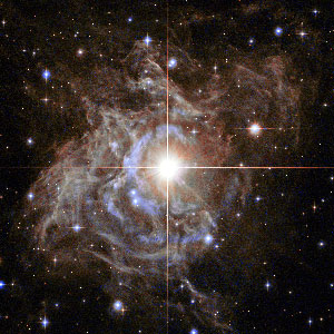 Lede art for "A Radically Conservative Solution for Cosmology’s Biggest Mystery": This Hubble image shows RS Puppis, a type of variable star known as a Cepheid variable. As variable stars go, Cepheids have comparatively long periods— RS Puppis, for example, varies in brightness by almost a factor of five every 40 or so days. RS Puppis is unusual; this variable star is shrouded by thick, dark clouds of dust enabling a phenomenon known as a light echo to be shown with stunning clarity. These Hubble observations show the ethereal object embedded in its dusty environment, set against a dark sky filled with background galaxies.