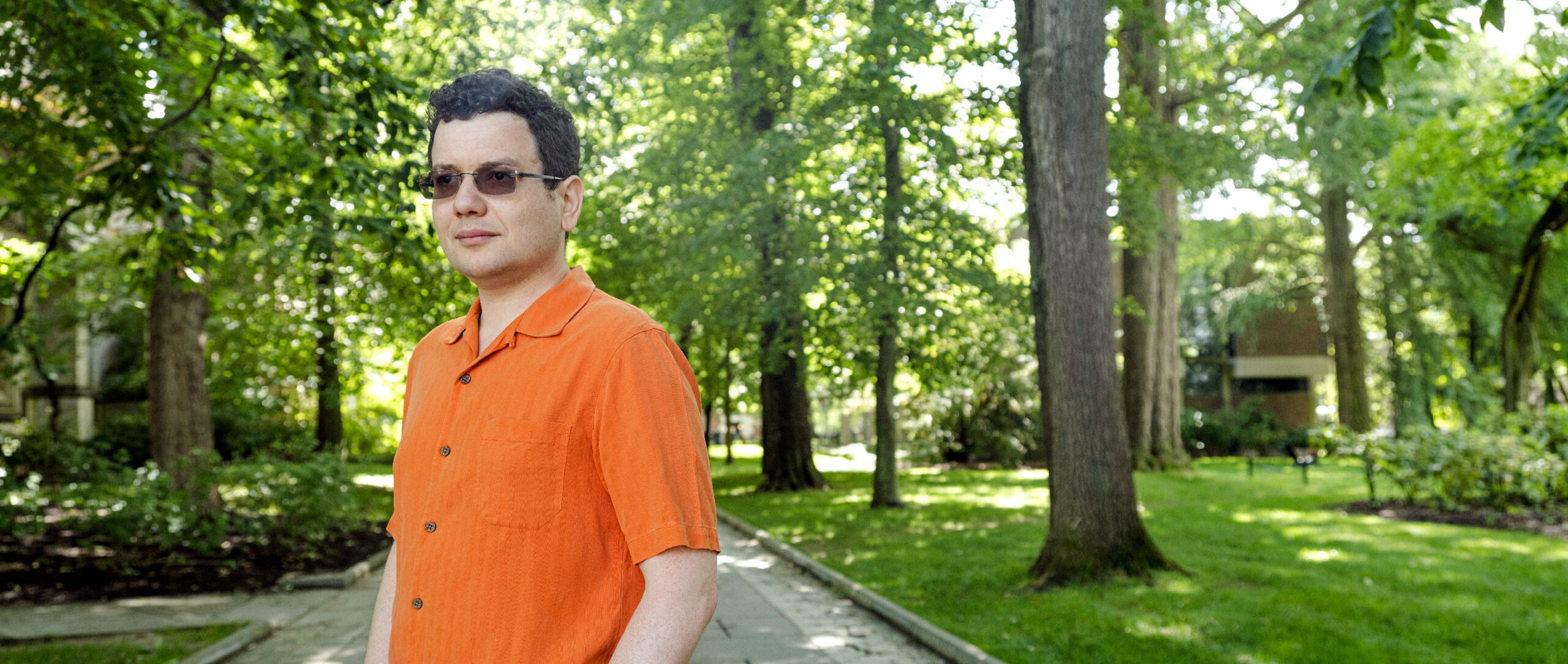 Mark Braverman, in an orange shirt, stands on a path lined with trees.