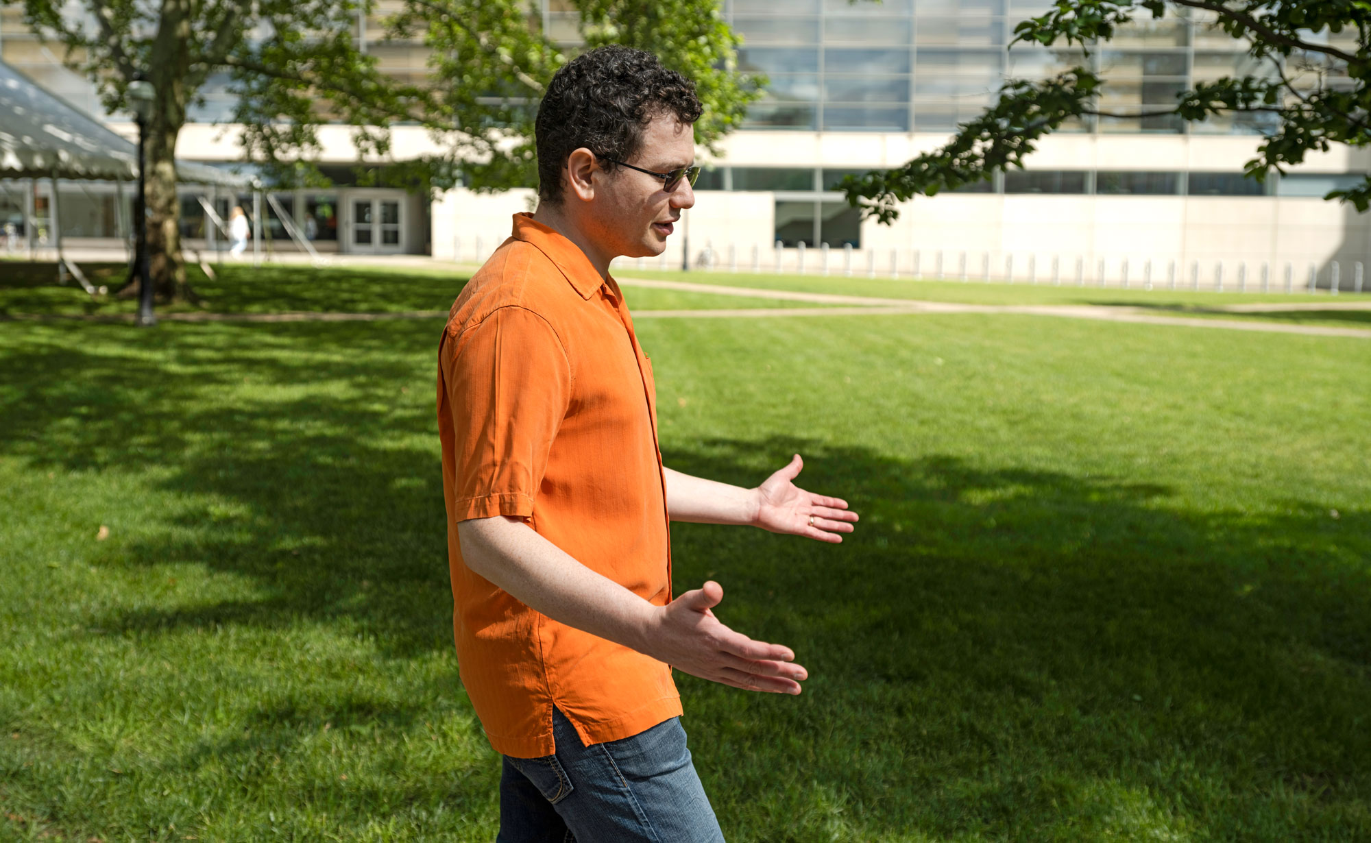 Mark Braverman, in an orange shirt, stands with his hands outstretched in a grassy field.