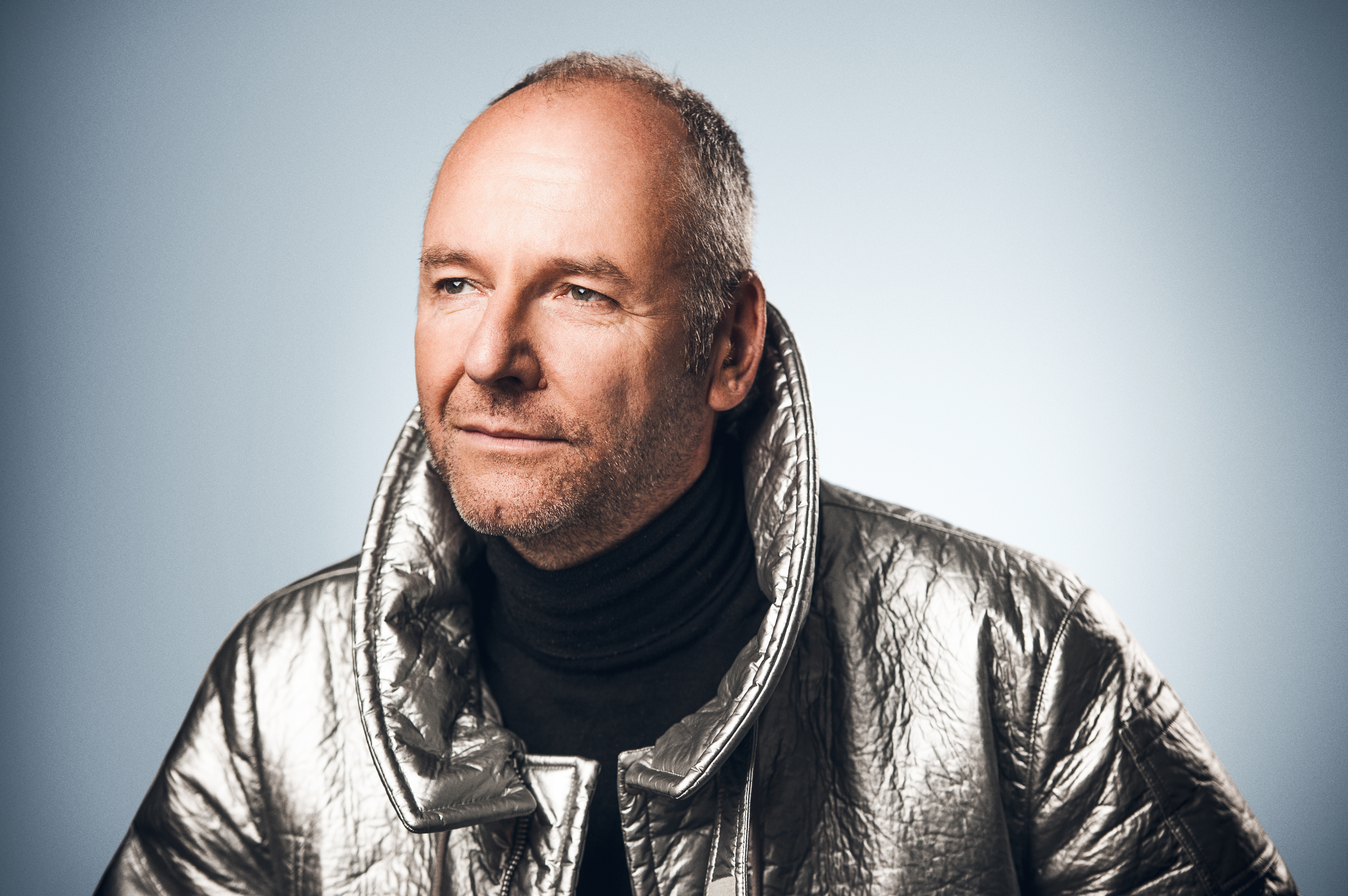 Photo of a smiling caucasian man wearing a silver jacket, with a light blue background.