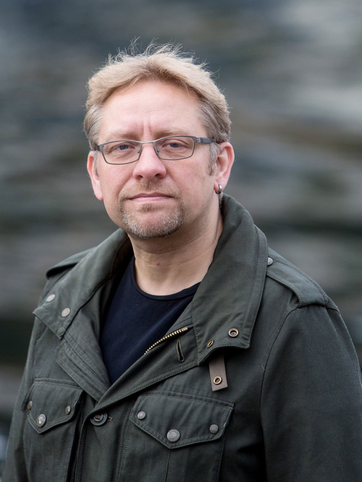 Photo of a man with short light colored hair, thin wireframed glasses, a dark shirt with a dark green jacket.