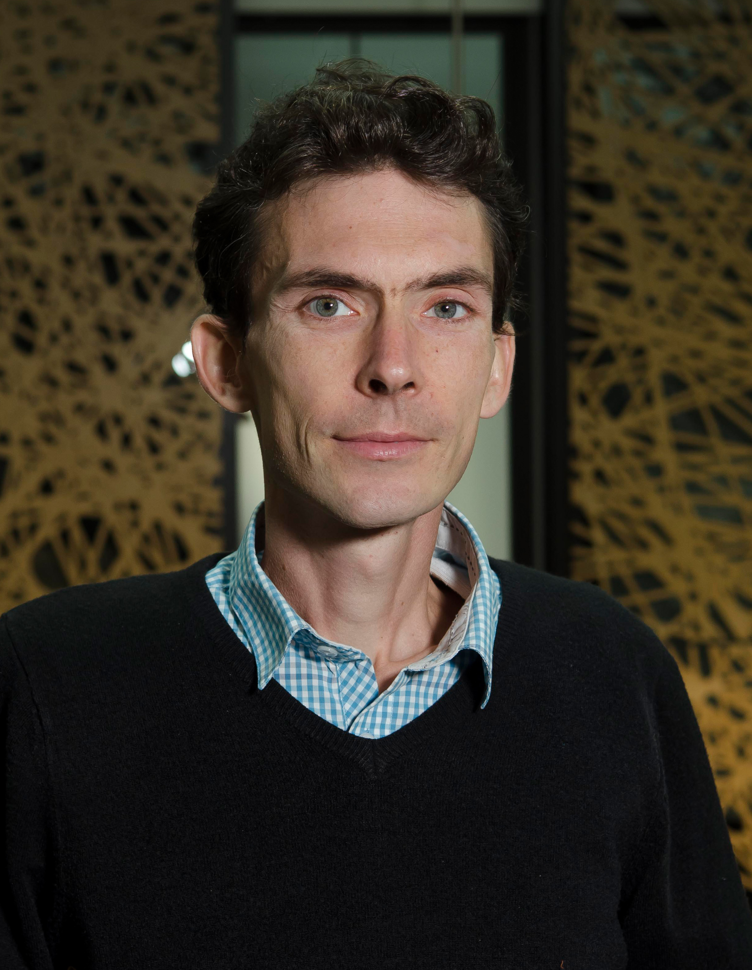 Photo of a man wearing a lightblue checkered collar shirt with a black jumper over it, looking into the camera.