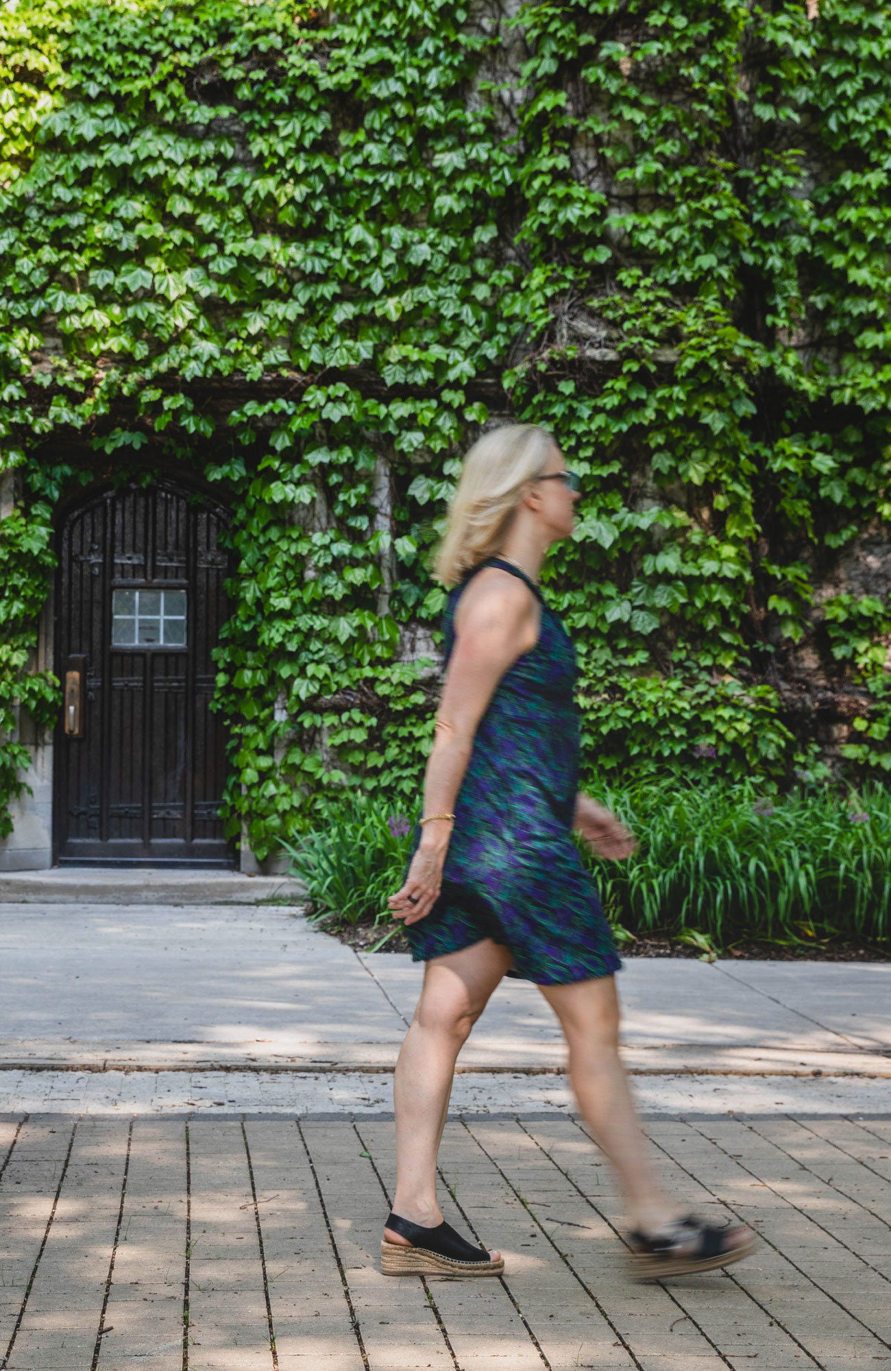 A woman with silver hair, walking in a blue dress with a wall of ivy in the background.