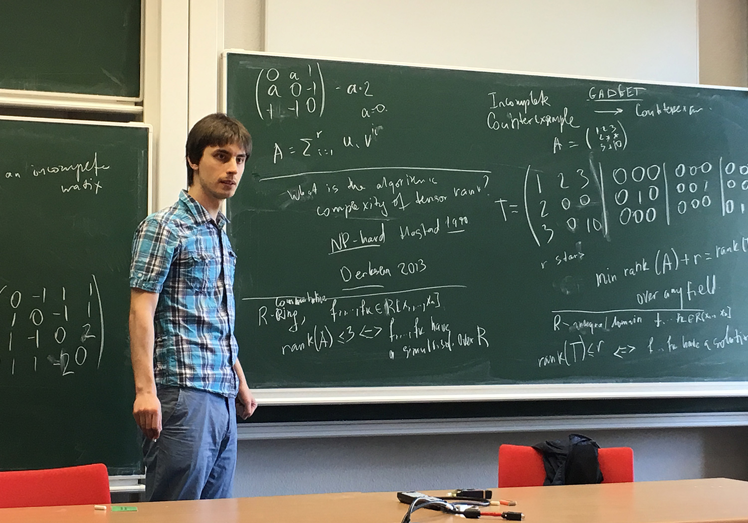 Photo of a caucasian man wearing a blue tartan shirt and blue slacks, holding a piece of chalk, standing next to a chalkboard in a classroom.