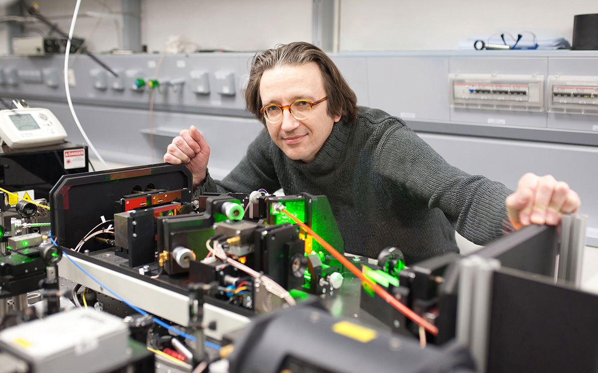 Photo of a man wearing glasses, a grey turtleneck longsleeve shirt, posing with a table top experiment setup with green lasers. He is smiling and looking into the camera.