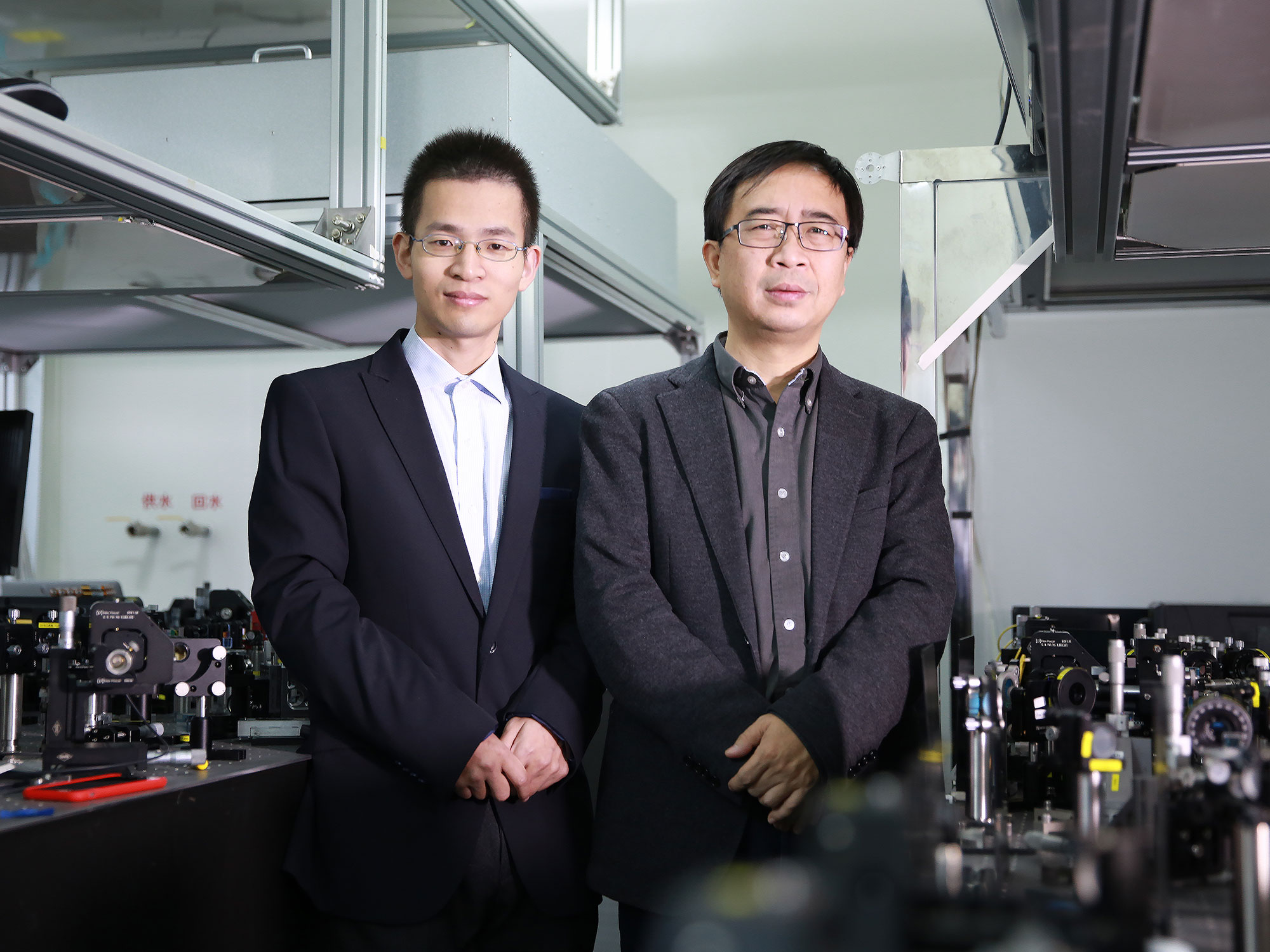 Photo of two men standing in a lab, both wearing glasses, button down shirts and suit jackets, arms resting on the front of their bodies with hands folded at waist level, looking into the camera.