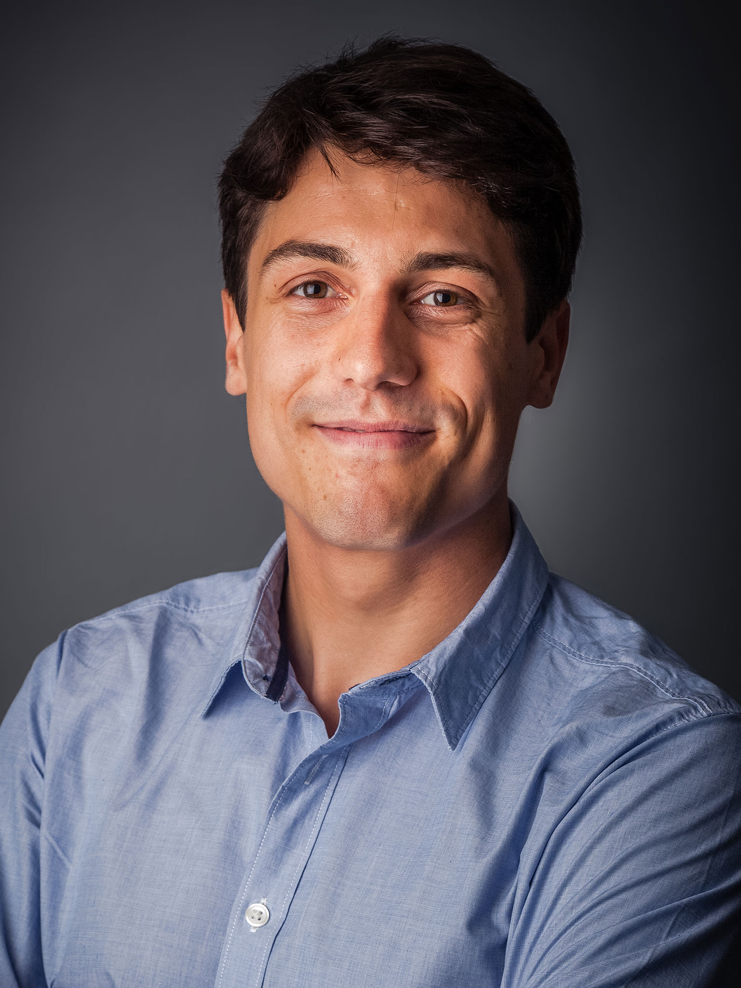 Studio portrait of a man wearing a light blue button down shirt, smiling with a grey background.