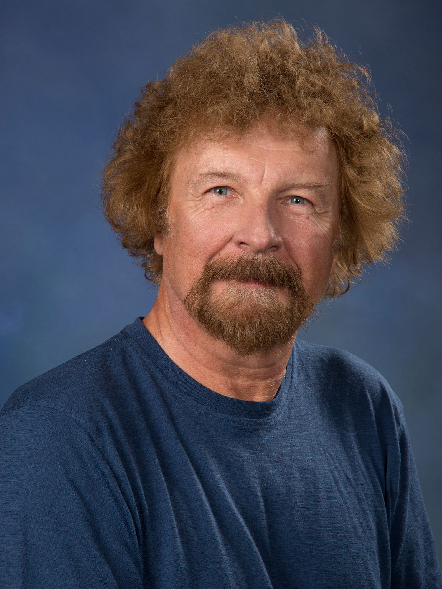 Photo os a man with a red trim beard and medium length red hair wearing a blue T-shirt looking into the camera against a blue background.