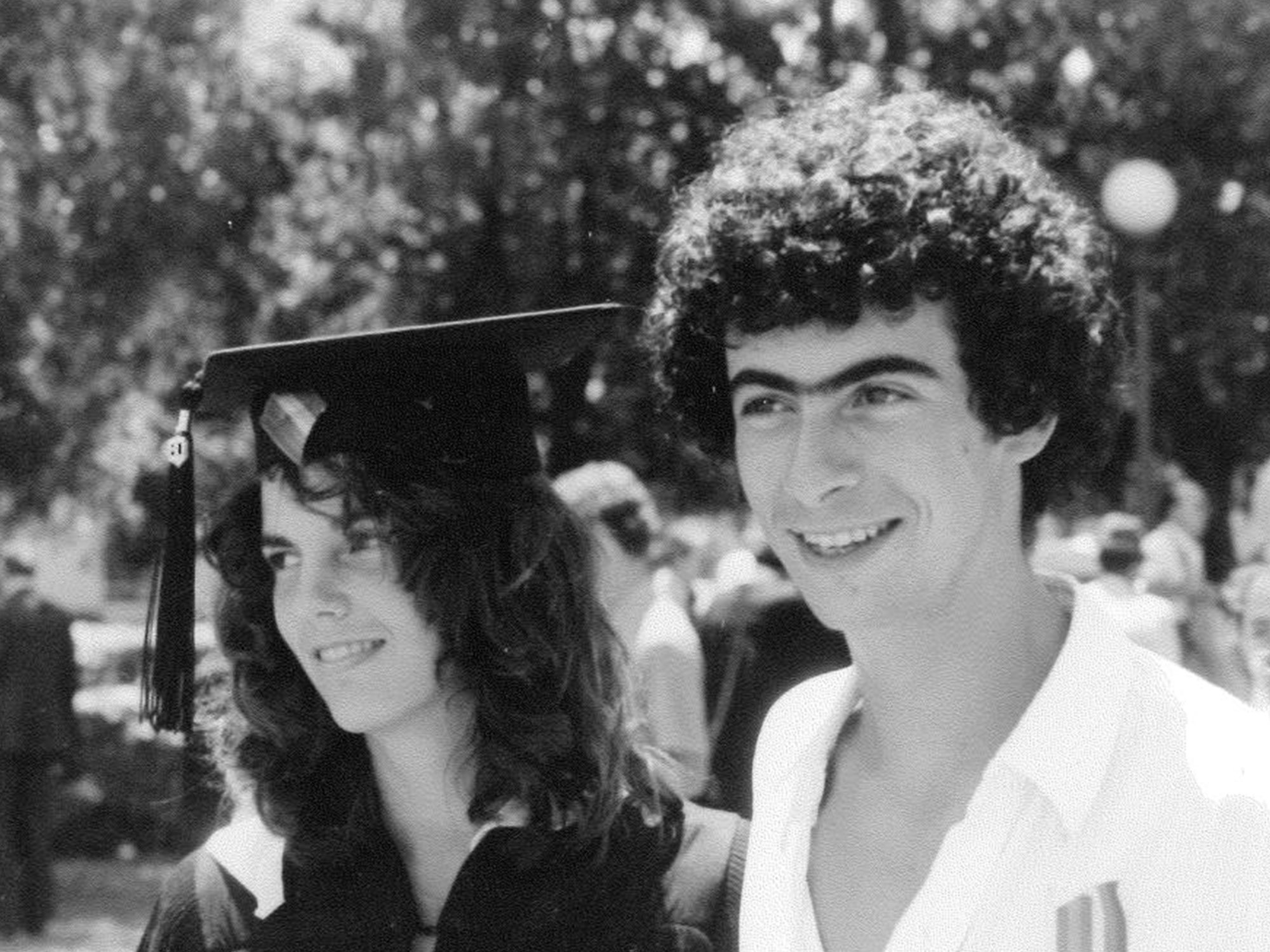 Black-and-white photo of Ann Nelson and David Kaplan standing side by side and smiling, with people and trees in the background. She is wearing a cap and gown; he is not.