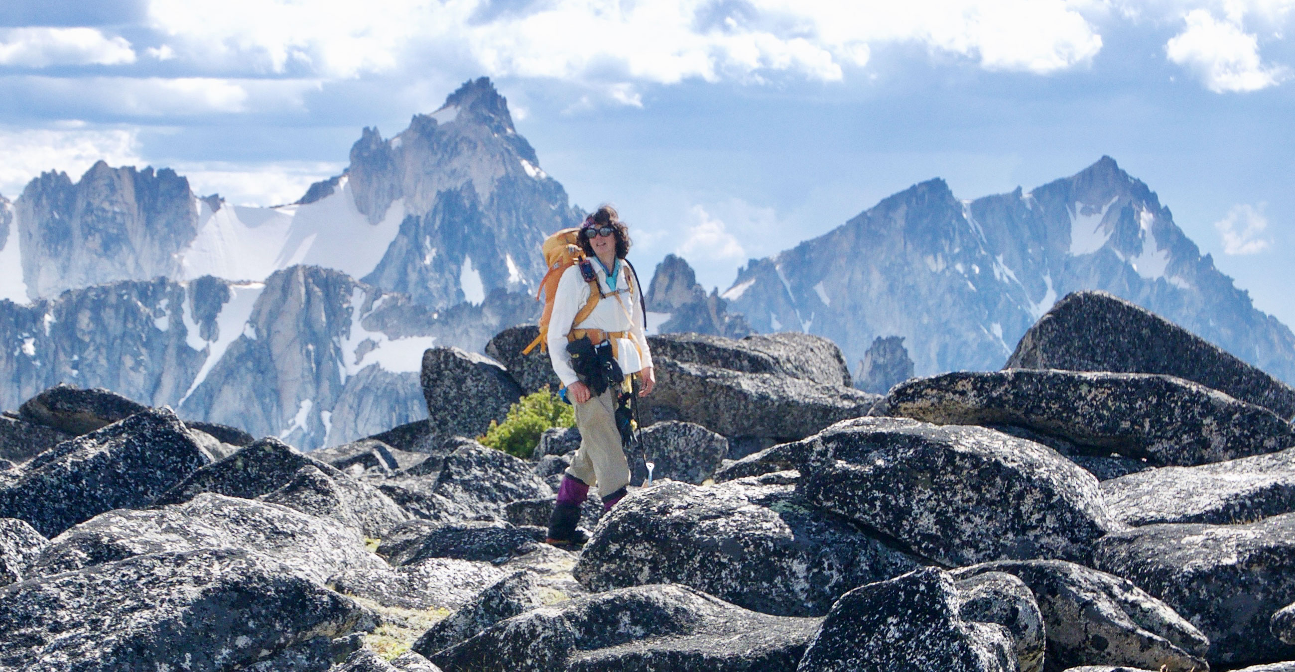 Ann Nelson in hiking gear traversing some boulders at the top of a mountain with snowy peaks in the background.