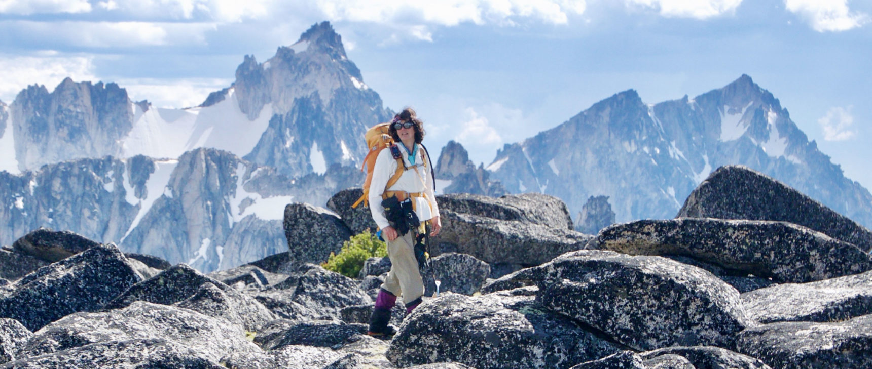 Ann Nelson in hiking gear traversing some boulders at the top of a mountain with snowy peaks in the background.