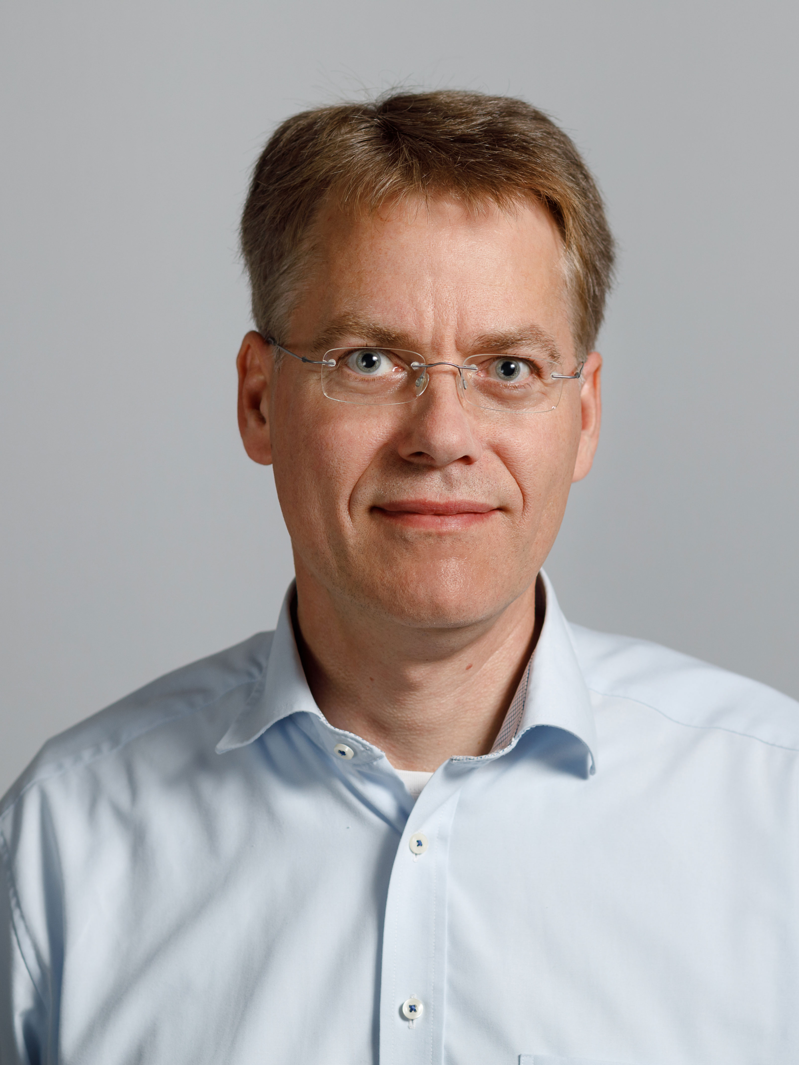 Portrait of a man with light colored eyes, wearing a pale blue button-down shirt, rimless glasses, smiling while looking straight into the camera.