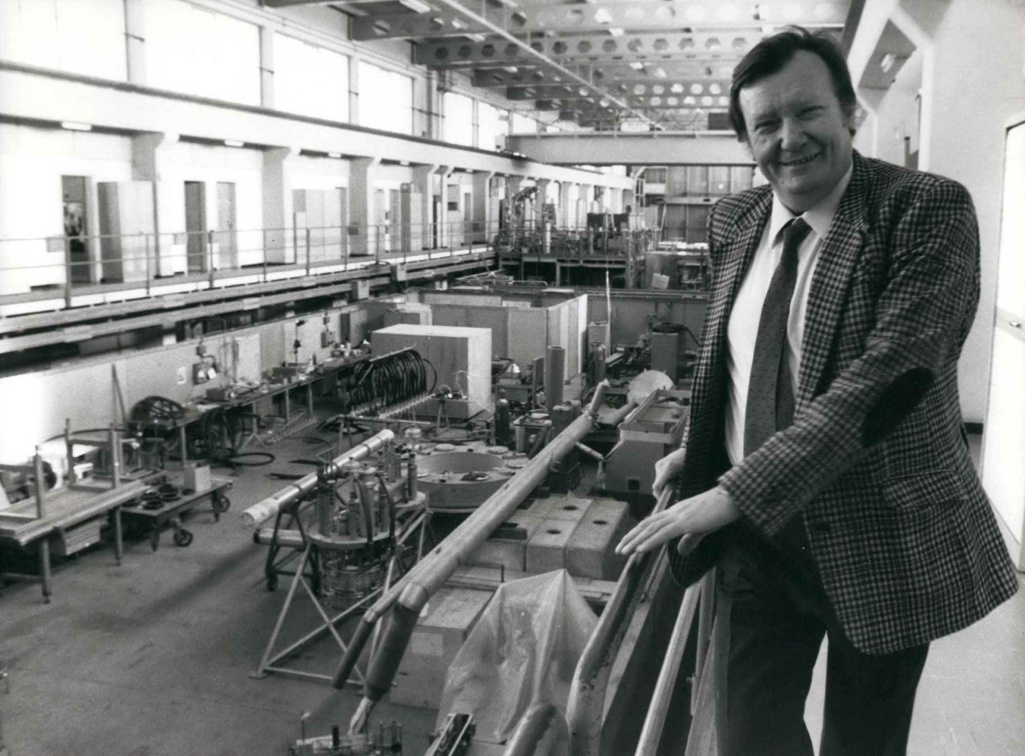 Black and white photo of a young Carlo Rubbia smiling in a suit and tie, standing on a walkway at CERN laboratory in 1984 with equipment behind him.