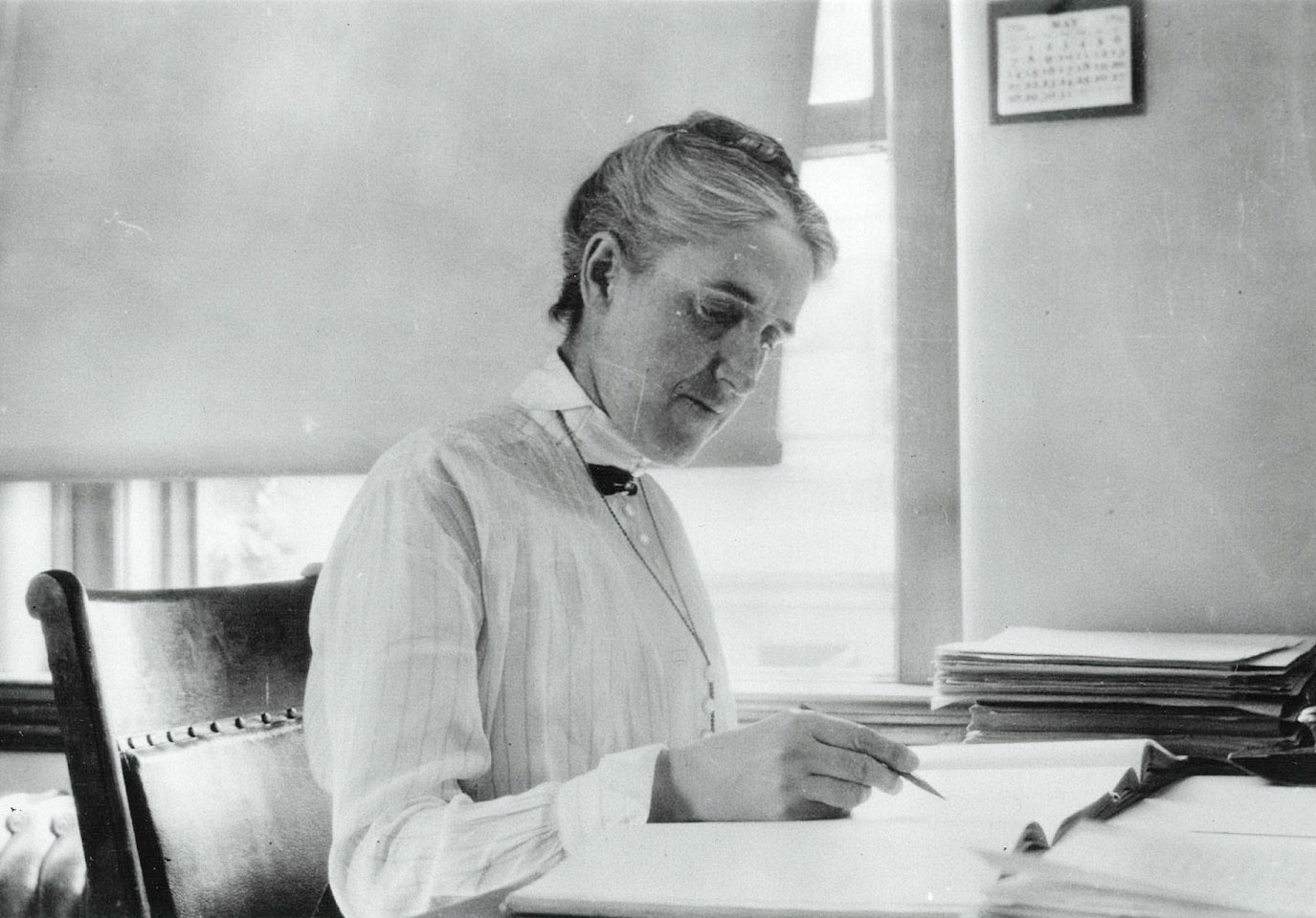 Black and white photo of Henrietta Swan Leavitt sitting at her desk near a window, holding a pen or pencil poised over some papers.