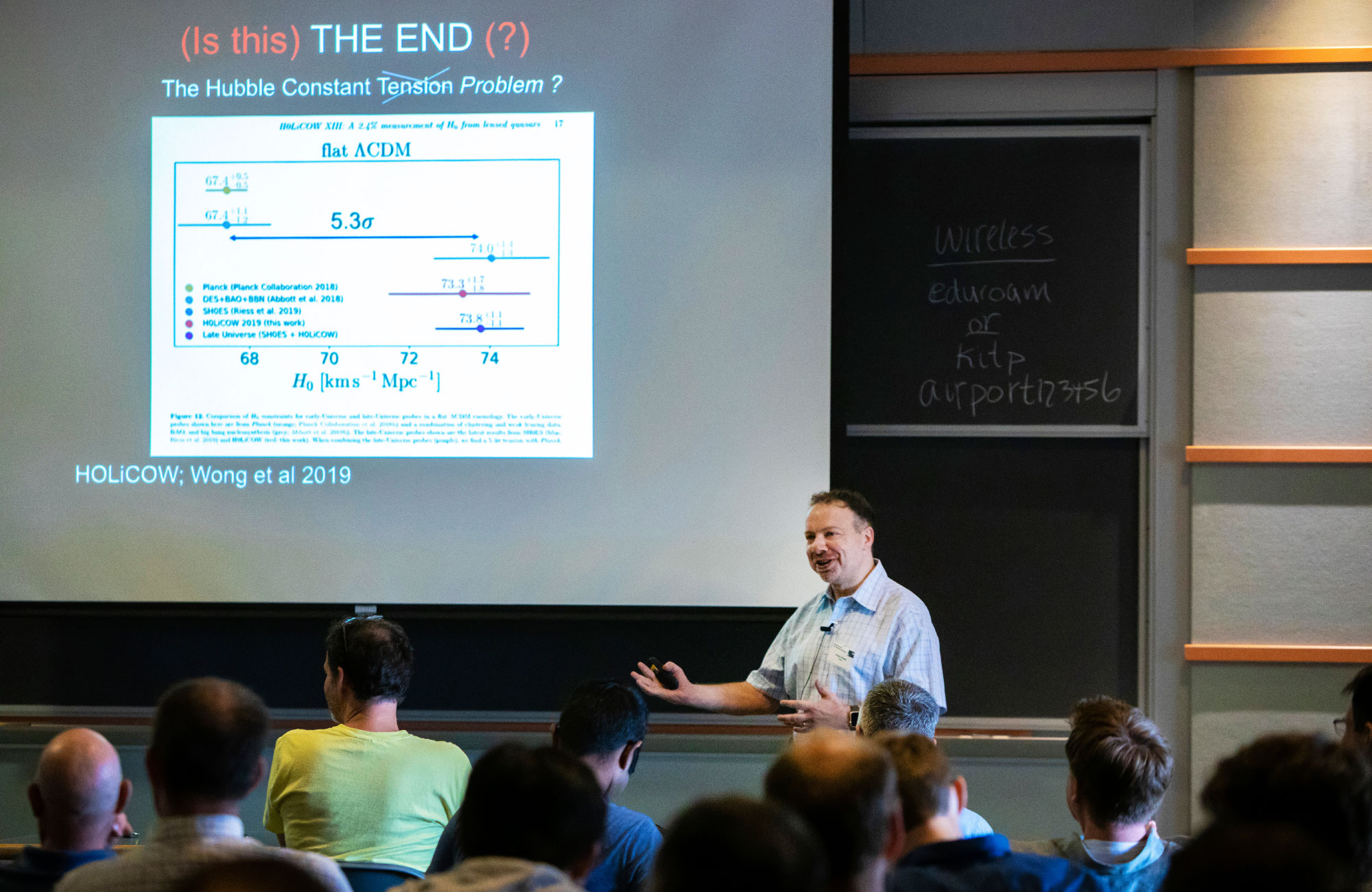 Adam Riess is smiling and gesturing to an audience of cosmologists with a slide on a screen behind him showing the discrepancy between predictions and observations of the cosmic expansion rate.