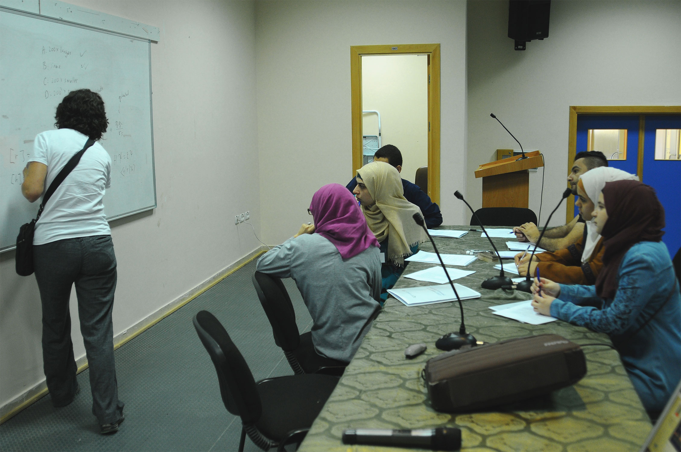 Ann Nelson writing at a dry-erase board in a classroom as four women in head scarves and two men look on.