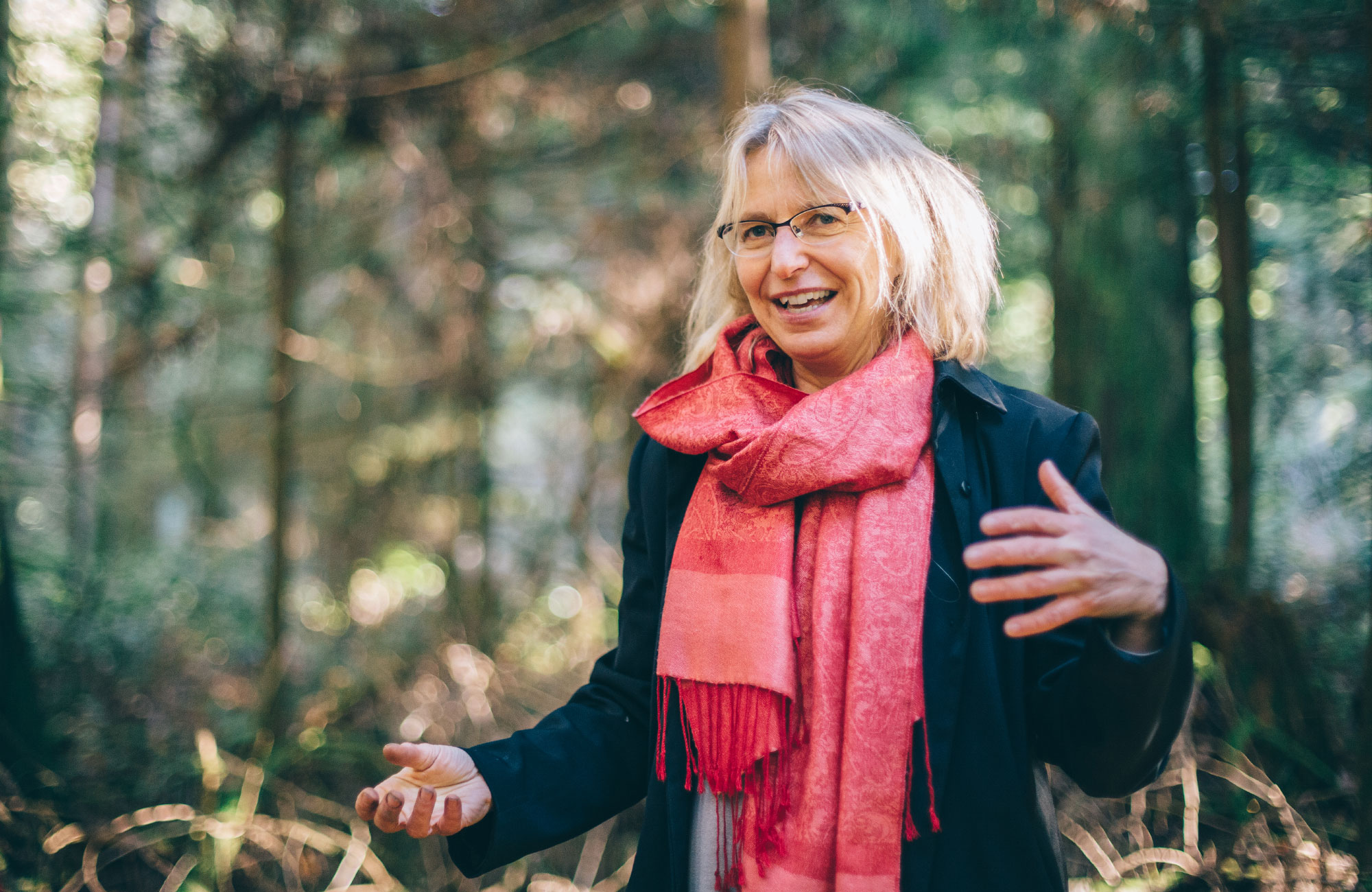 Suzanne Simard, wearing a dark coat and rose-colored scarf, stands in a forest.