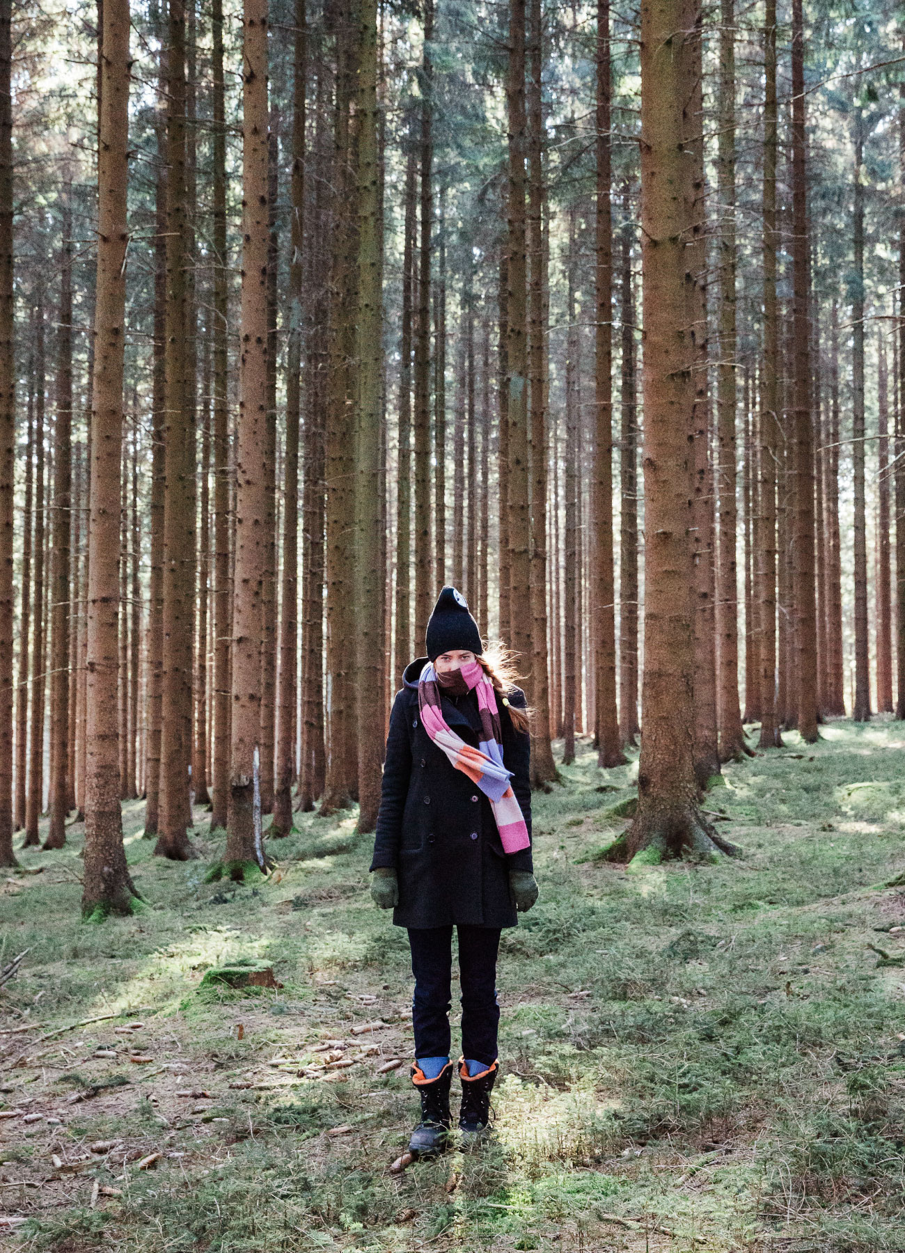 Toby Kiers, in a dark coat, dark winter hat and long striped scarf, stands in a forest of tall spruce trees.