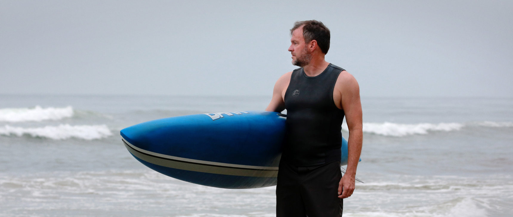 Craig Callender holding a paddleboard at the beach.