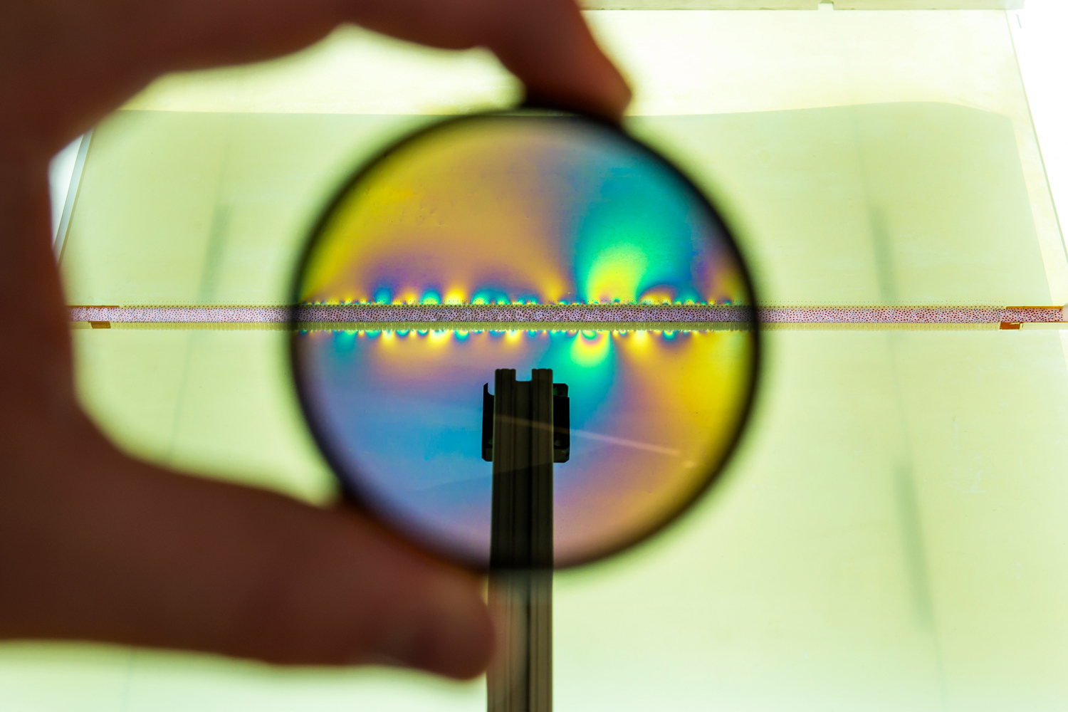 A hand holds a lens in front of the seam between two plastic blocks. Through the lens, colorful light emanates from stress points in the seam.