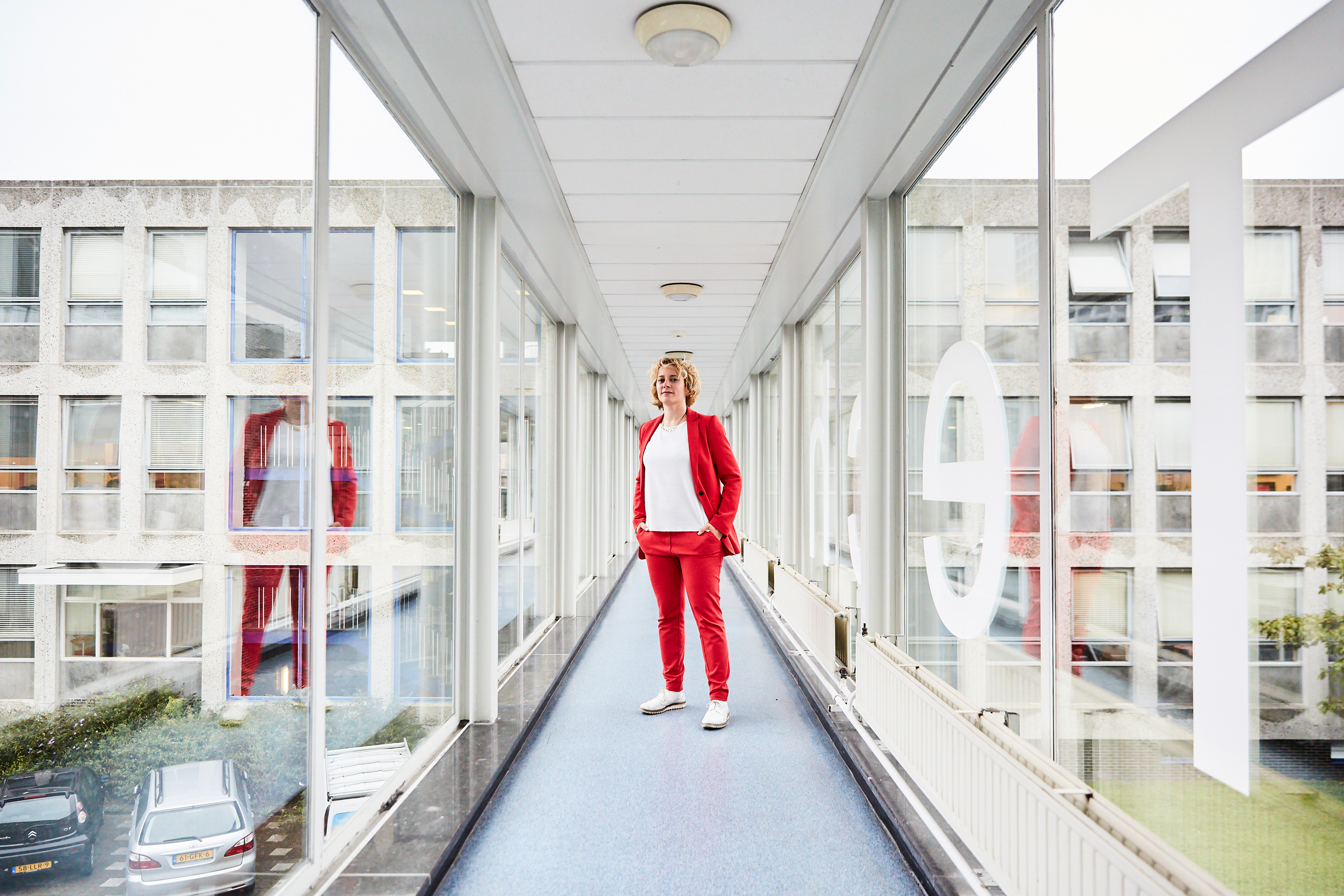 Stephanie Wehner in a red suit standing in a glass-paneled corridor at Delft University of Technology in the Netherlands. Her reflection appears in the glass to the right and left.]