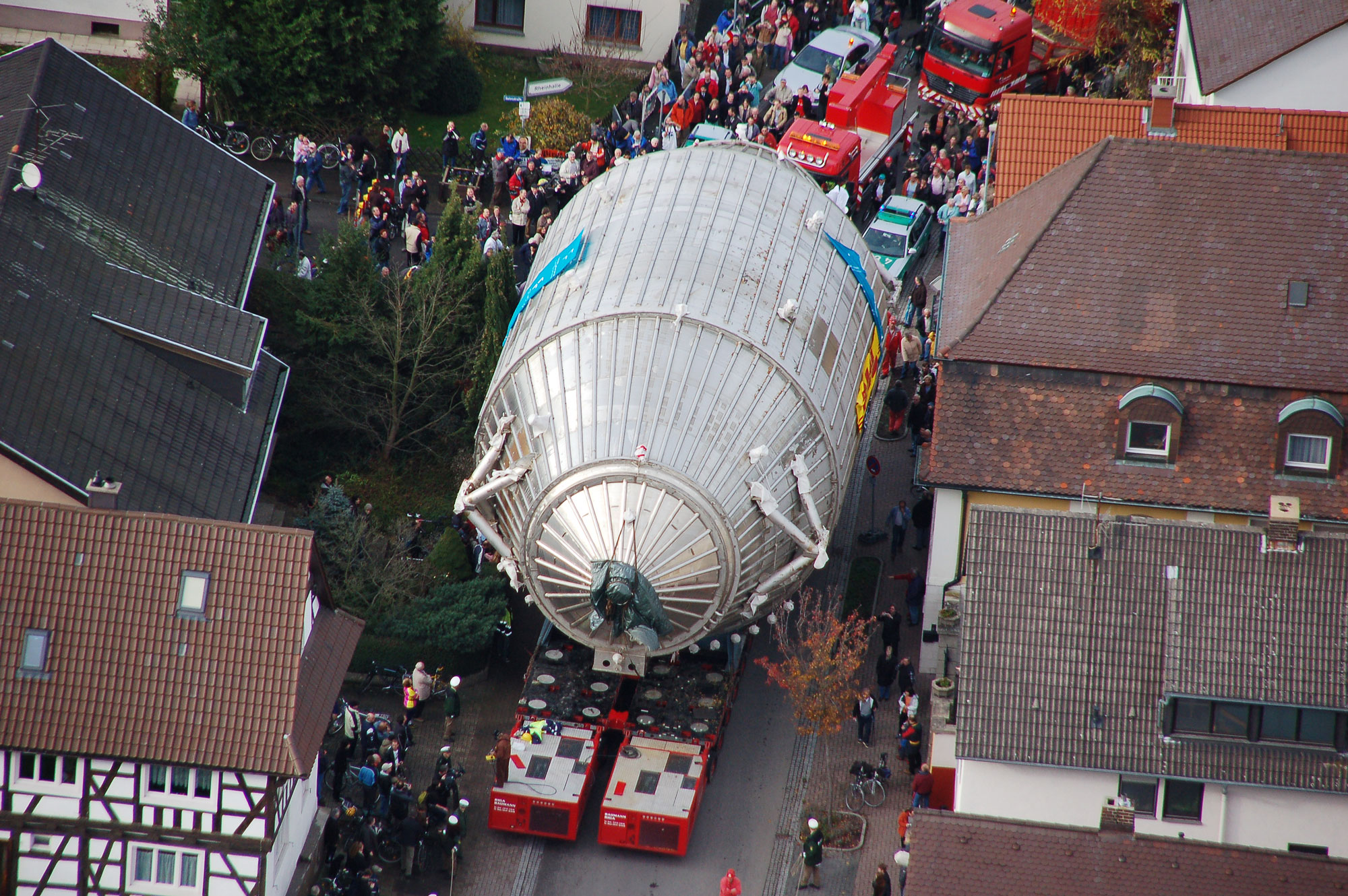 A large, egg-shaped metallic structure is seen from above as red emergency vehicles transport it through a quaint European village.