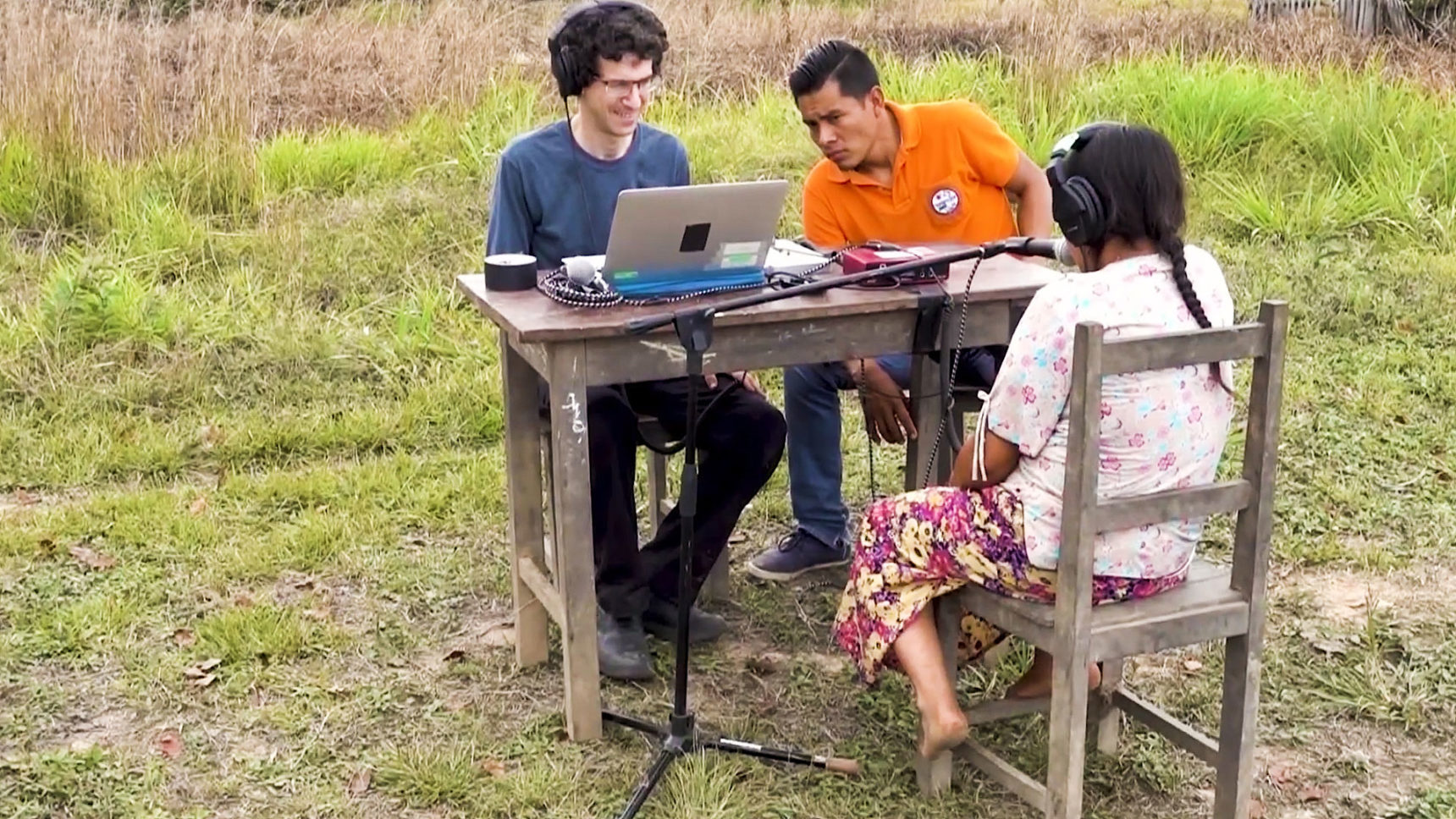 At a table in the Bolivian rainforest, a Tsimané volunteer wears headphones and sits at a microphone, while a scientist and interpreter observe the test.