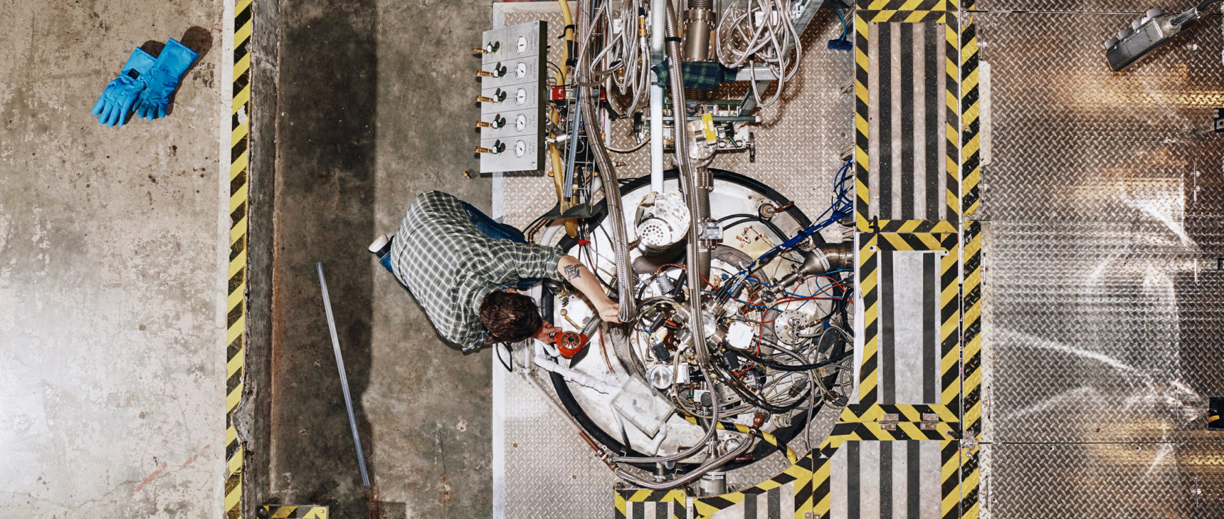 An overhead shot of an engineer working on a metal tank with pipes and cables coming out of it.