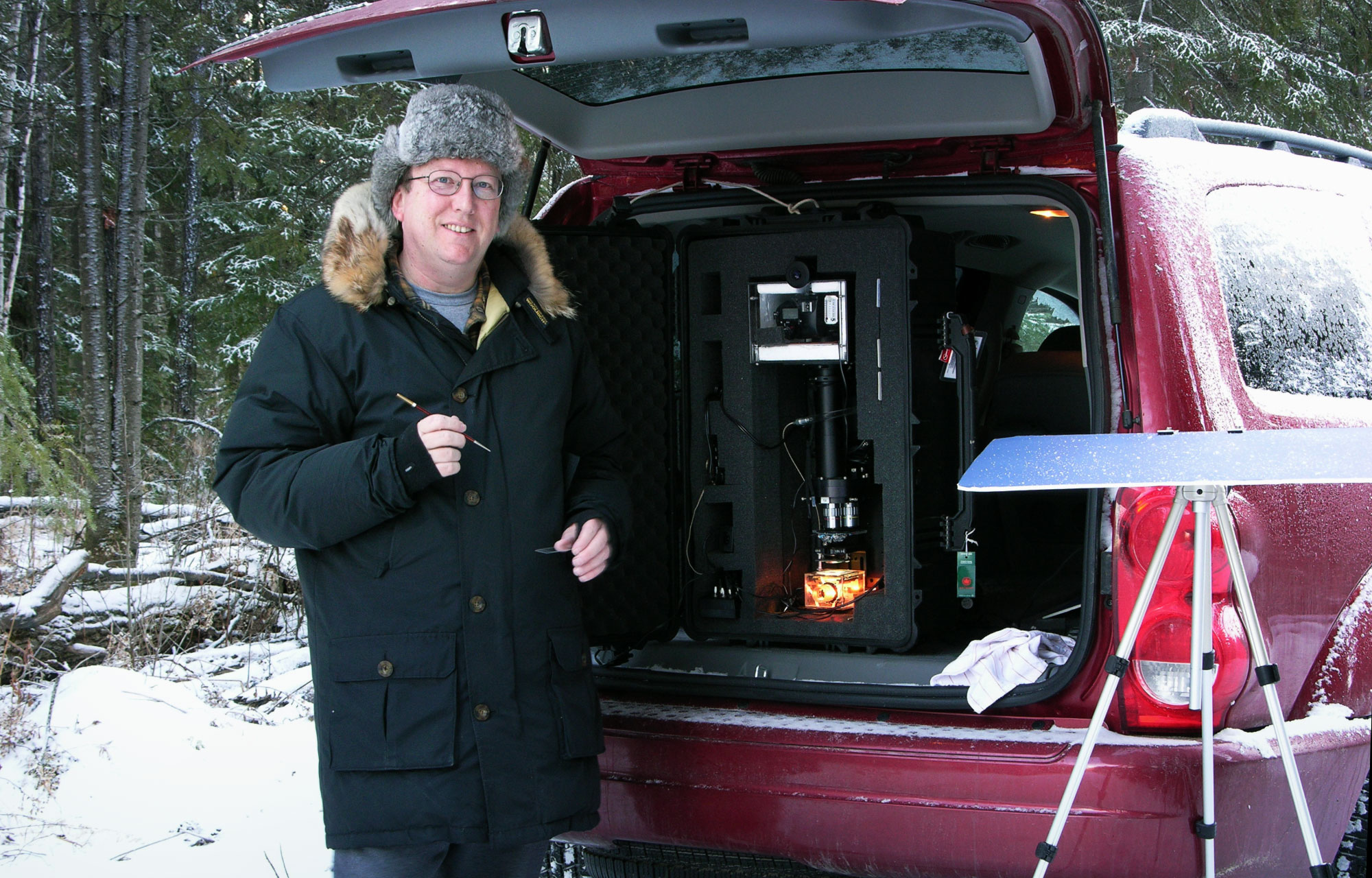 Kenneth Libbrecht, smiling in a parka, stands next to his vehicle in a snowy wooded area. A foam board rests on a tripod and a microscope is visible in the trunk of the car.