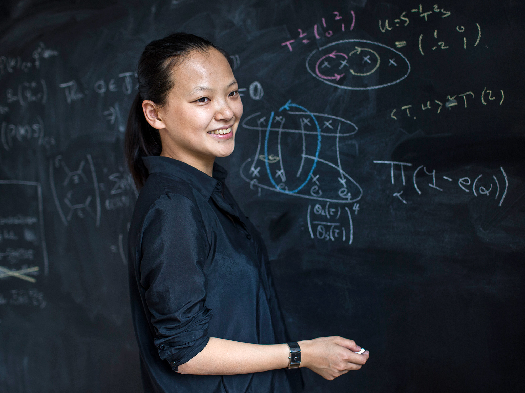 Miranda Cheng shown standing in front of a chalkboard that's covered in calculations, holding a piece of chalk.