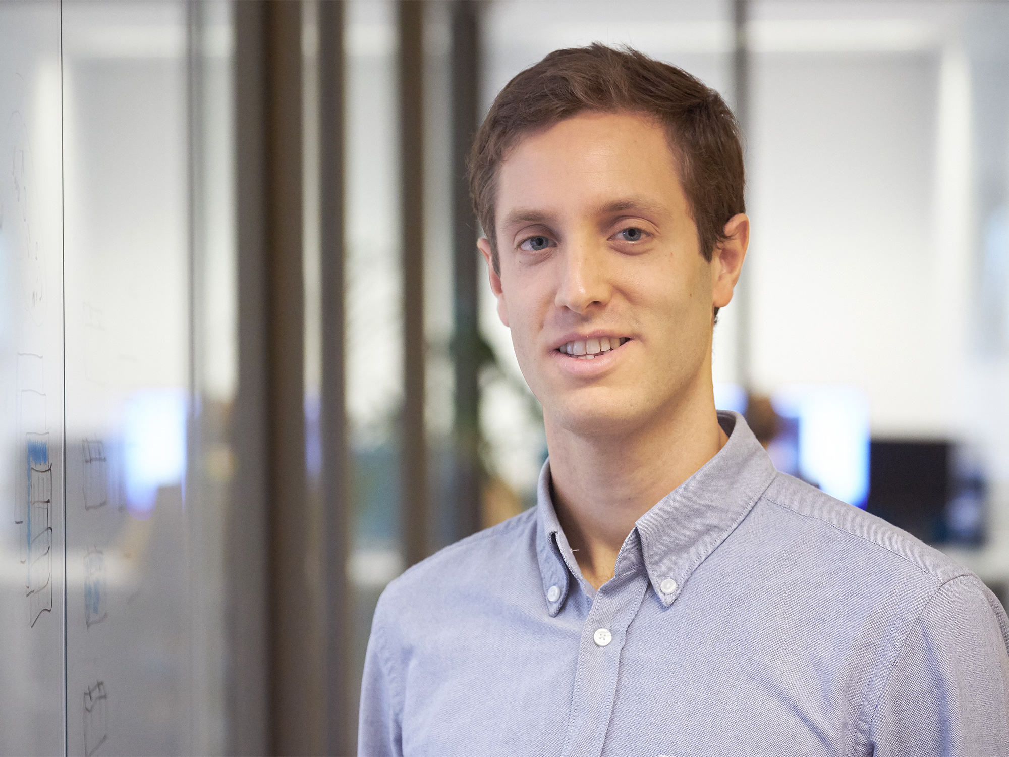 Taco Cohen, wearing a blue Oxford shirt and standing next to a dry-erase board, smiles at the camera.