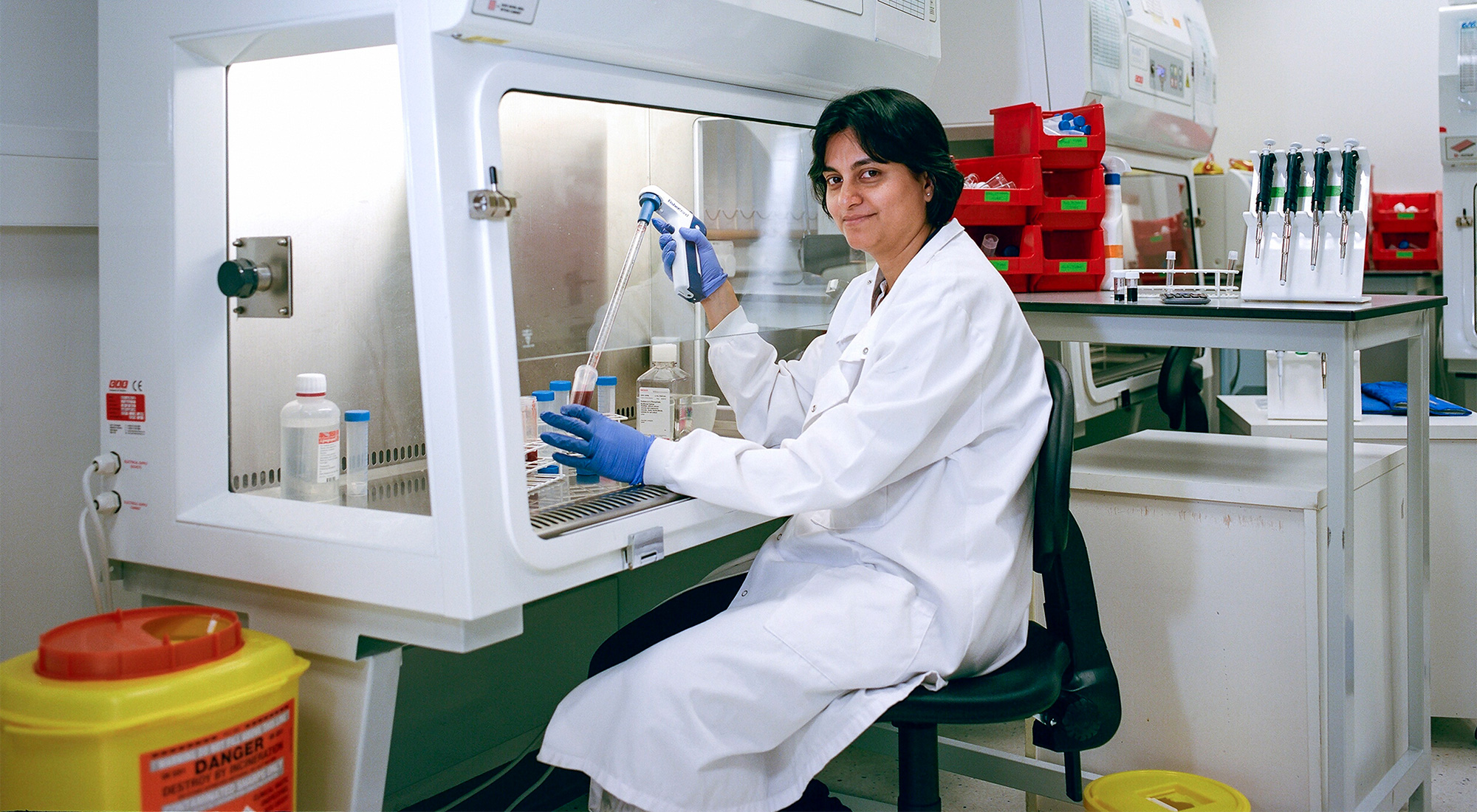 Muzlifah Haniffa sits at a sterile hood, pipetting blood from a tube..