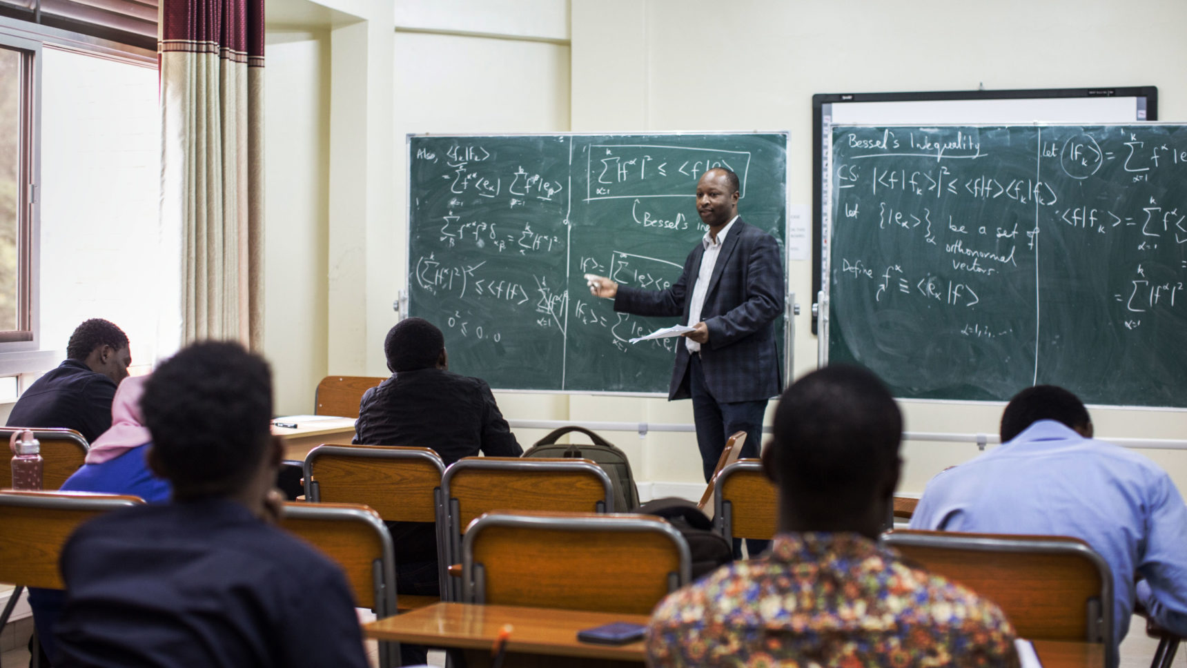 Omololu Akin-Ojo in a blue checked suit standing at the front of a class, gesturing toward equations on a green chalkboard.