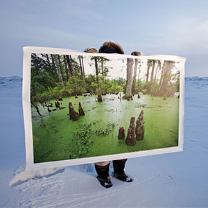 A man standing in the Arctic tundra holds up a large photo of a swamp.