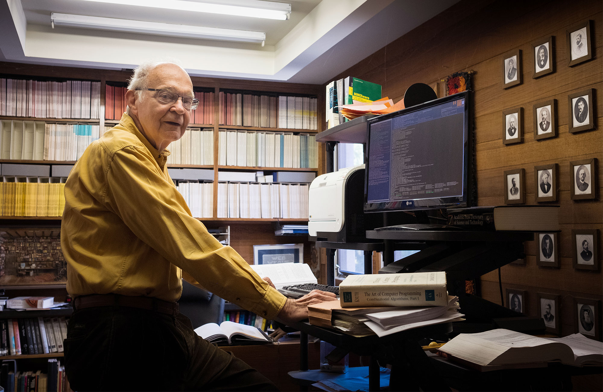 Photo of Donald Knuth at a computer in his home, in front of shelves of books and a wall full of portraits