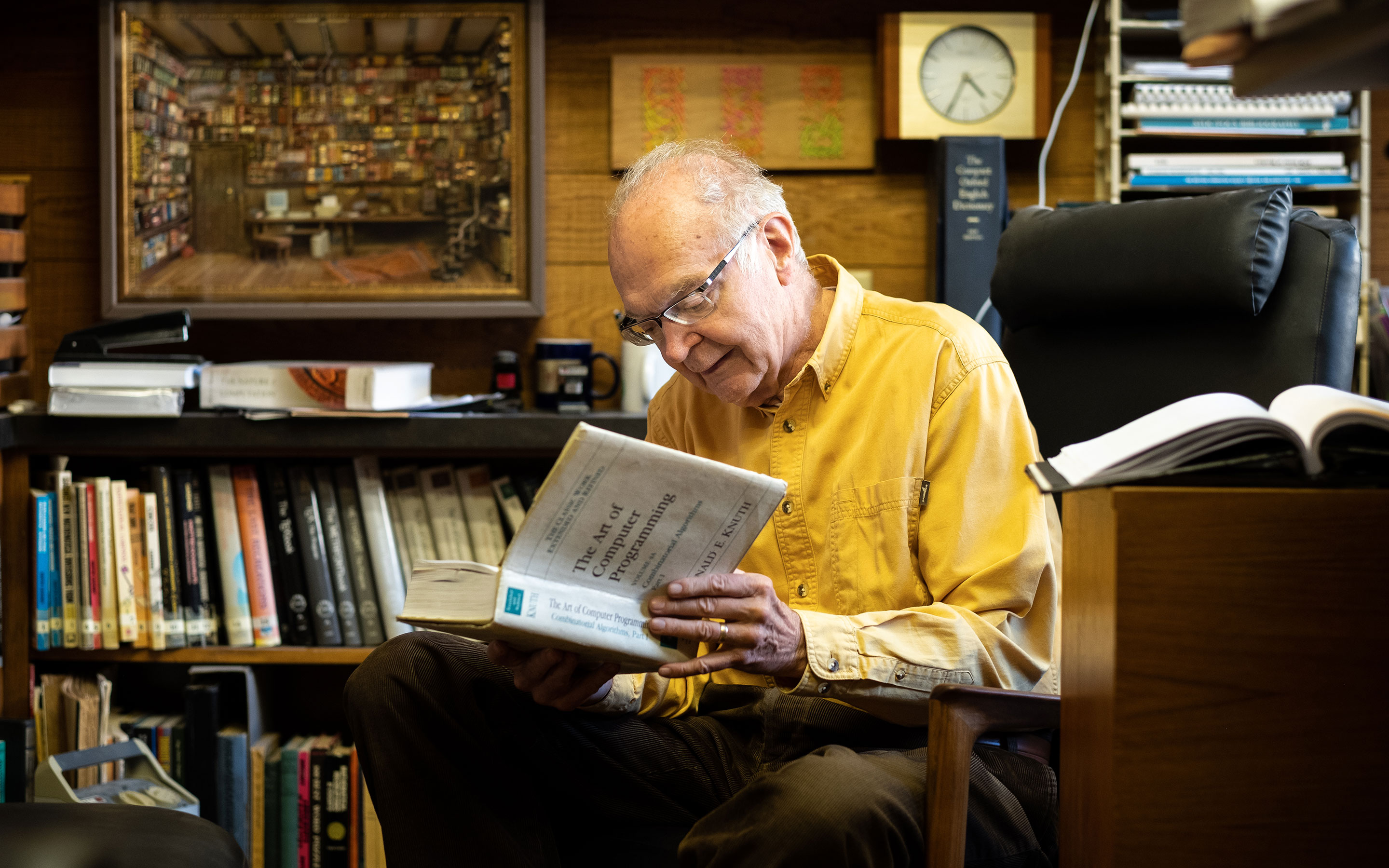 Photo of Donald Knuth in a yellow shirt sits and reads from a volume of The Art of Computer Programming