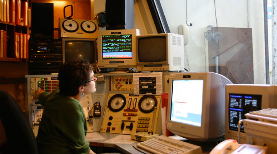 Photo of Emily Levesque sitting at a control panel for a variety of electronics and computers.