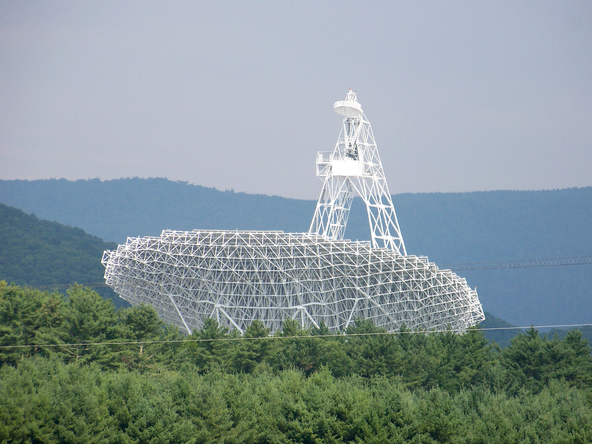 The large dish of a radio telescope pointed toward the sky against a forest landscape.