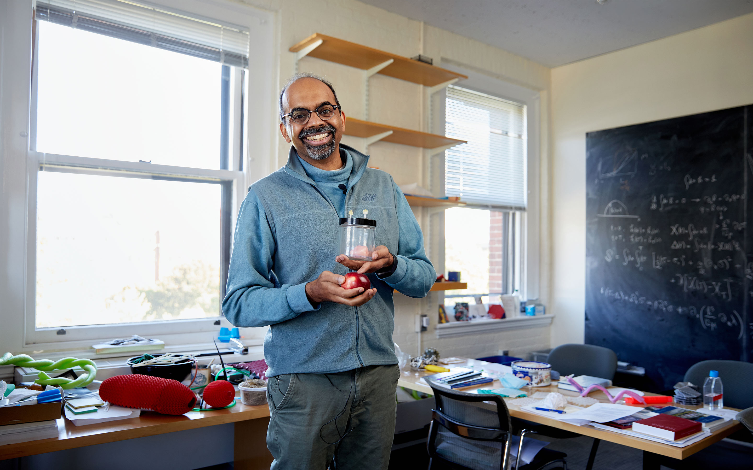 Harvard professor L. Mahadevan stands in a slightly cluttered office, holding an apple and a jar containing a miniature brain