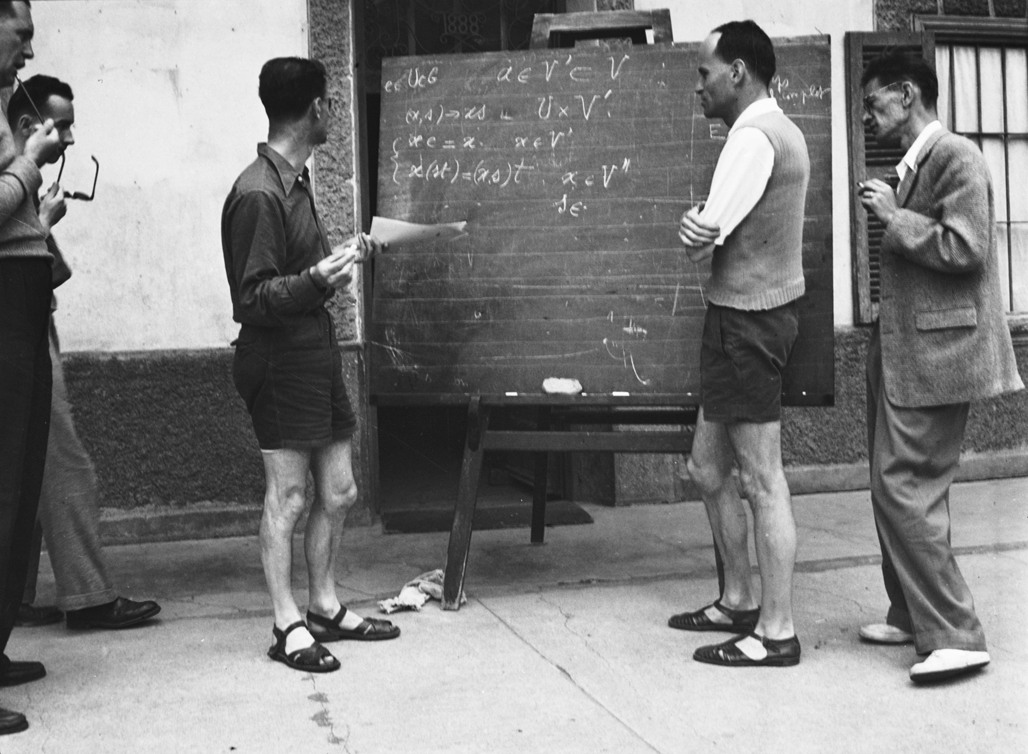 Black and white historical photo of five men standing around a chalkboard outside a building