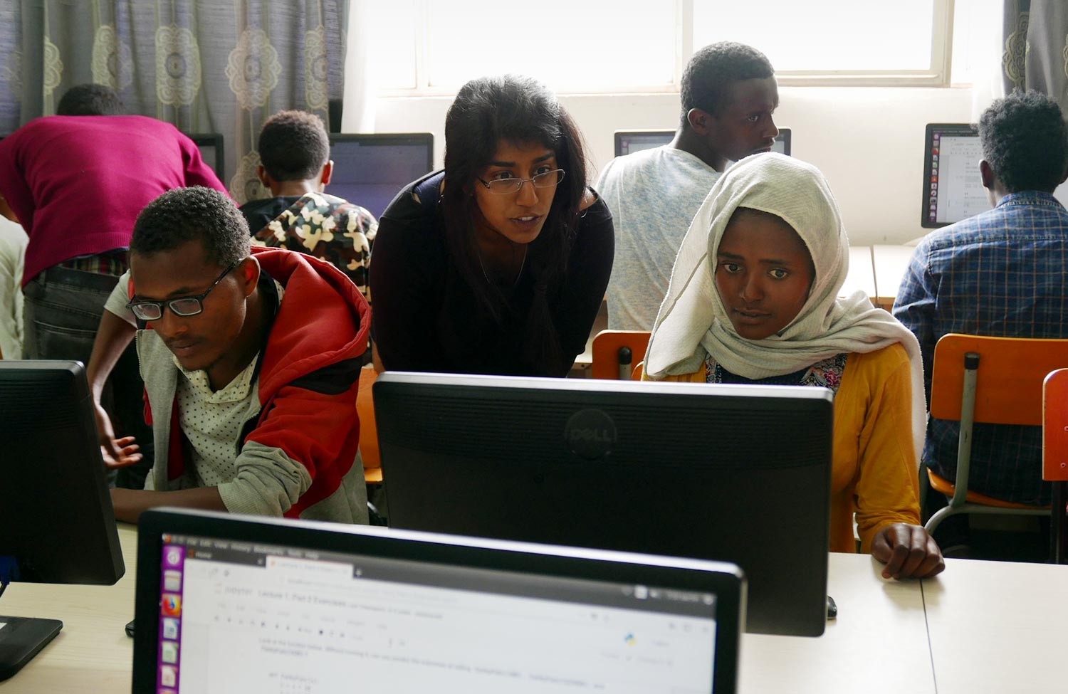 A diptych. Left image is a group shot of participants in the AddisCoder program. Right image is in an AddisCoder classroom, where a teacher is leaning over a student’s shoulder while the student works at a computer.