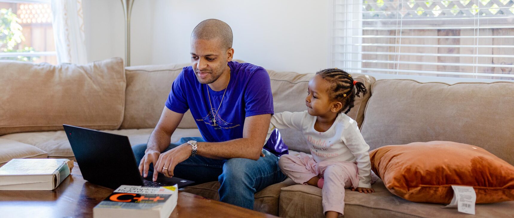 Computer scientist Jelani Nelson seated on a couch next to his daughter. There are textbooks and a laptop on the coffee table in front of him.