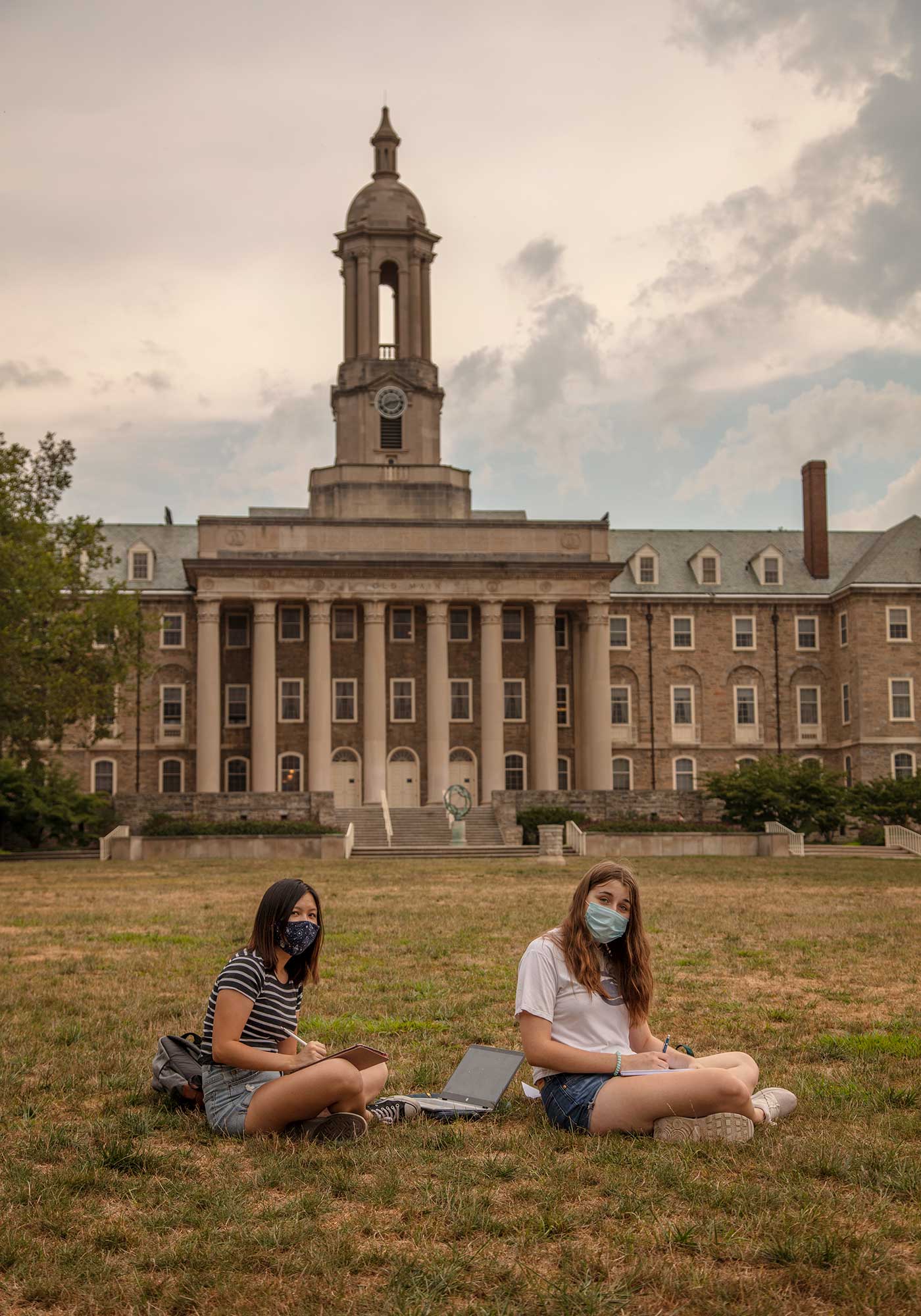 Two students wearing masks sit on a lawn at Pennsylvania State University.