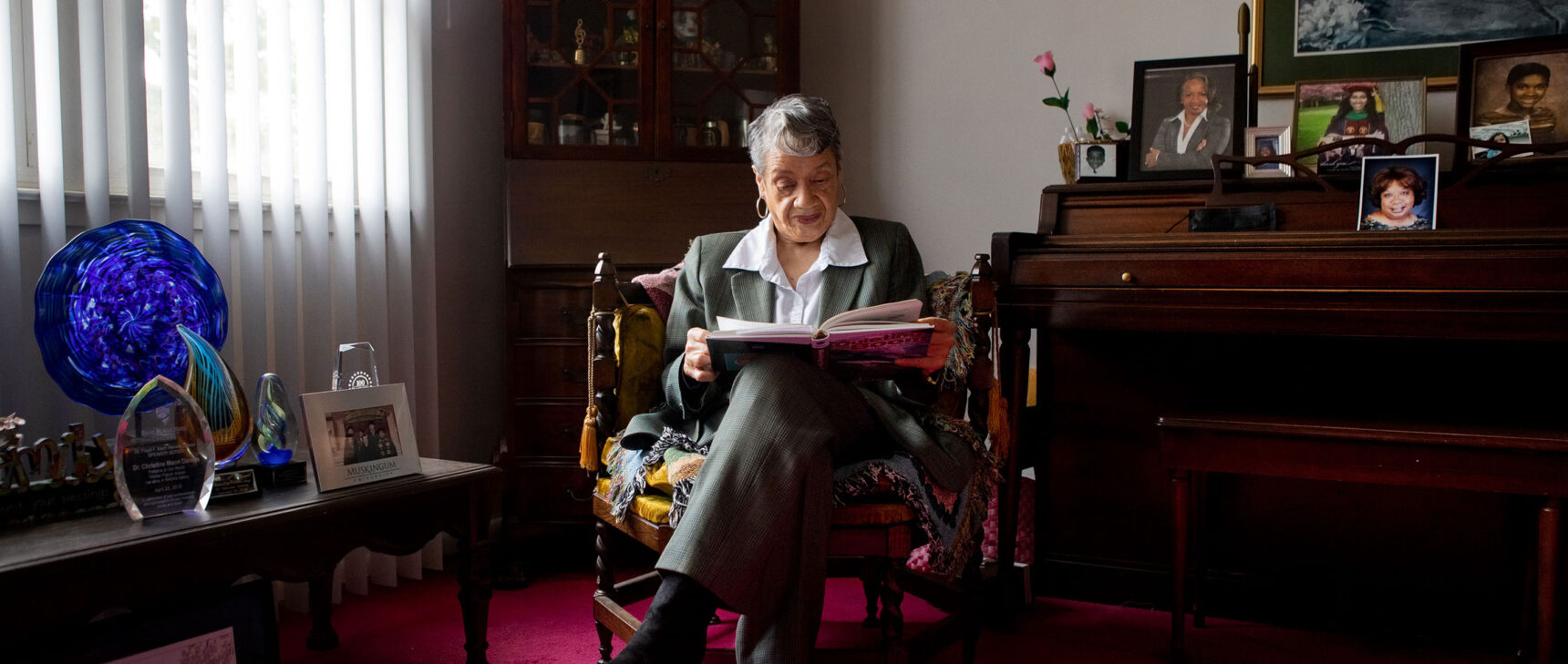 Photo of Christine Darden in a gray suit, seated reading a book in her home