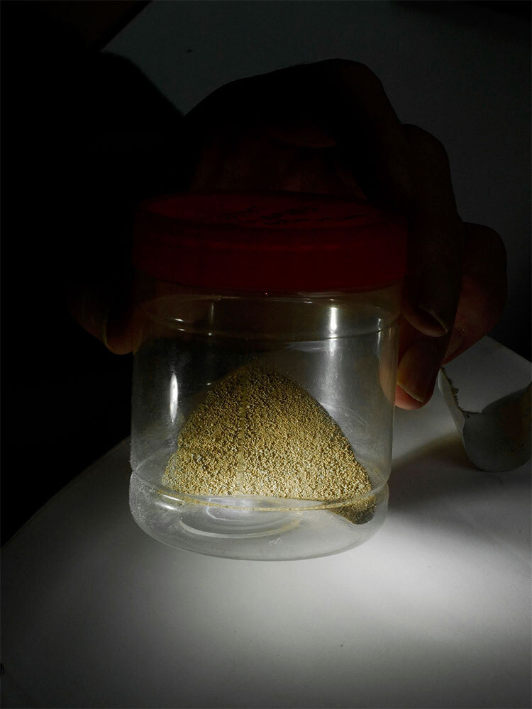 A hand holds up a jar of tawny-colored dust against a black background.