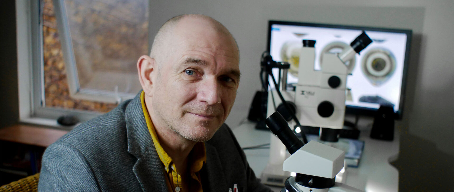 Matthew Genge in a yellow shirt and gray jacket seated at a microscope.